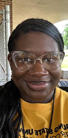 Close-up outdoor portrait of a person wearing large clear-framed eyeglasses and a yellow T-shirt. The person is facing the camera beneath a covered outdoor walkway. A brick column, chain-link fence, and greenery are visible in the background. Partially visible black text appears on the T-shirt. The image is framed closely around the face and shoulders and lit with natural daylight.