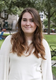 Outdoor portrait of a person with long brown hair wearing a light-colored blouse and cardigan. The person is standing and facing the camera in a park-like setting with trees, grass, benches, and campus-style buildings visible in the background. Natural daylight provides soft, even lighting.
