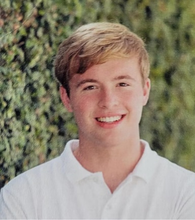 Outdoor portrait of a person with short, light-colored hair wearing a white collared polo shirt. The person is facing the camera with greenery and trees softly blurred in the background. The image is framed from the chest up and lit by natural daylight.