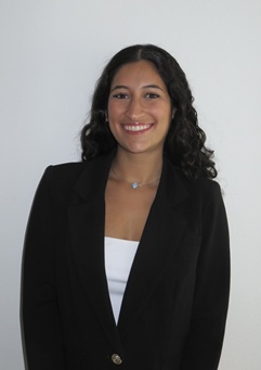 Head-and-shoulders portrait of a person with shoulder-length dark curly hair wearing a black blazer over a white top. The person is facing the camera against a plain light gray background. A small pendant necklace is visible, and the image is evenly lit with studio-style lighting.