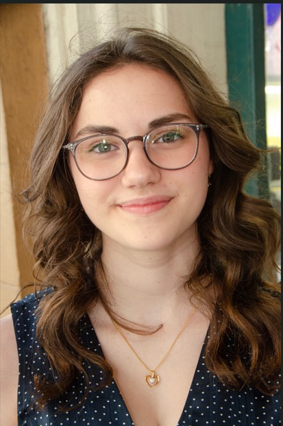 Outdoor portrait of a person with shoulder-length wavy hair wearing rectangular eyeglasses and a sleeveless dark blue dress with a small white dot pattern. A necklace with a heart-shaped pendant is visible. The person is facing the camera in front of a building entrance with wooden beams and windows in the background. The image is framed from the chest up and lit with soft natural daylight.