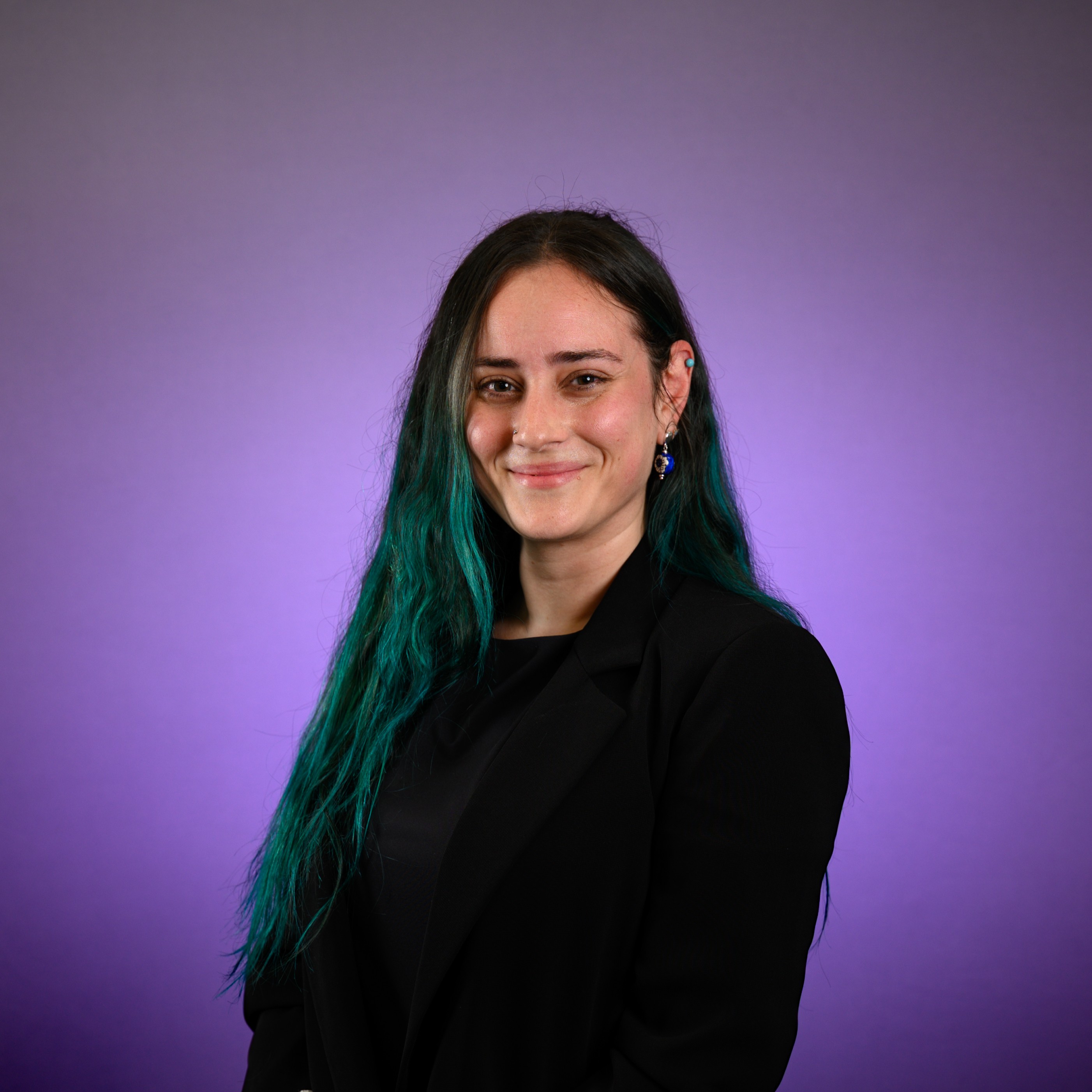 Portrait of a person with long dark hair featuring teal-colored ends, wearing a black blazer and dangling earrings, facing the camera against a soft purple gradient background.