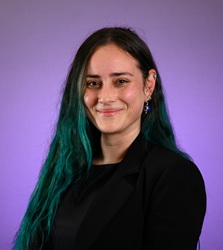Portrait of a person with long dark hair featuring teal-colored ends, wearing a black blazer and dangling earrings, facing the camera against a soft purple gradient background.