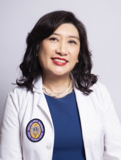 Professional studio portrait of a person seated against a light gray background. The person is wearing a white medical coat with a circular university-style emblem on the left side, a blue top, and a pearl necklace. The image is framed from the chest up, with dark shoulder-length hair and red lipstick. The portrait is evenly lit and presented in a clinical or academic professional style.