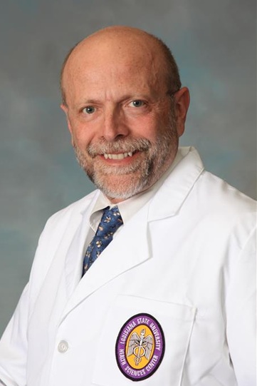 Professional studio portrait of a person wearing a white medical coat with a circular Louisiana State University Health Sciences Center emblem on the chest pocket and a blue patterned tie. The image is framed from the chest up against a neutral gray-blue background. The person has closely cropped hair and a neatly trimmed beard and mustache. The portrait is evenly lit and presented in a clinical or academic professional style.