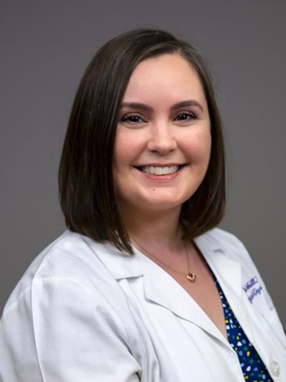 Professional head-and-shoulders portrait of a person wearing a white medical coat over a patterned blue top and a small pendant necklace. The image is framed from the shoulders up against a plain gray background. The person has shoulder-length dark hair and is facing the camera. The portrait is evenly lit and presented in a clinical or academic professional style.
