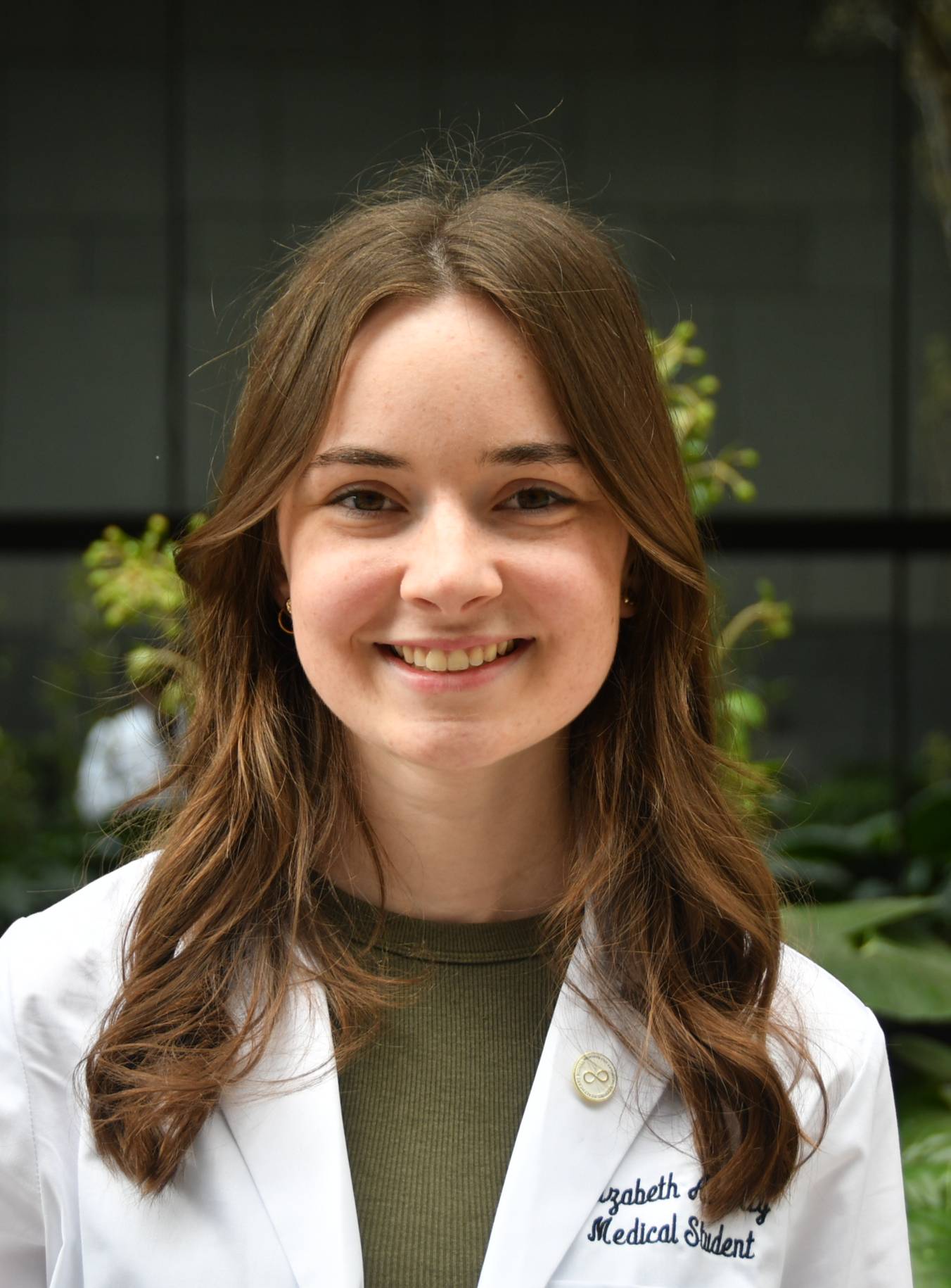 Professional medical school portrait showing a person wearing a white medical coat with embroidered text identifying the wearer as a medical student and a small lapel pin on the coat. The subject is standing outdoors among lush green plants, wearing an olive-green top beneath the coat. A modern glass building and additional greenery are visible in the background. The image is lit by natural daylight with the subject centered in the frame.
