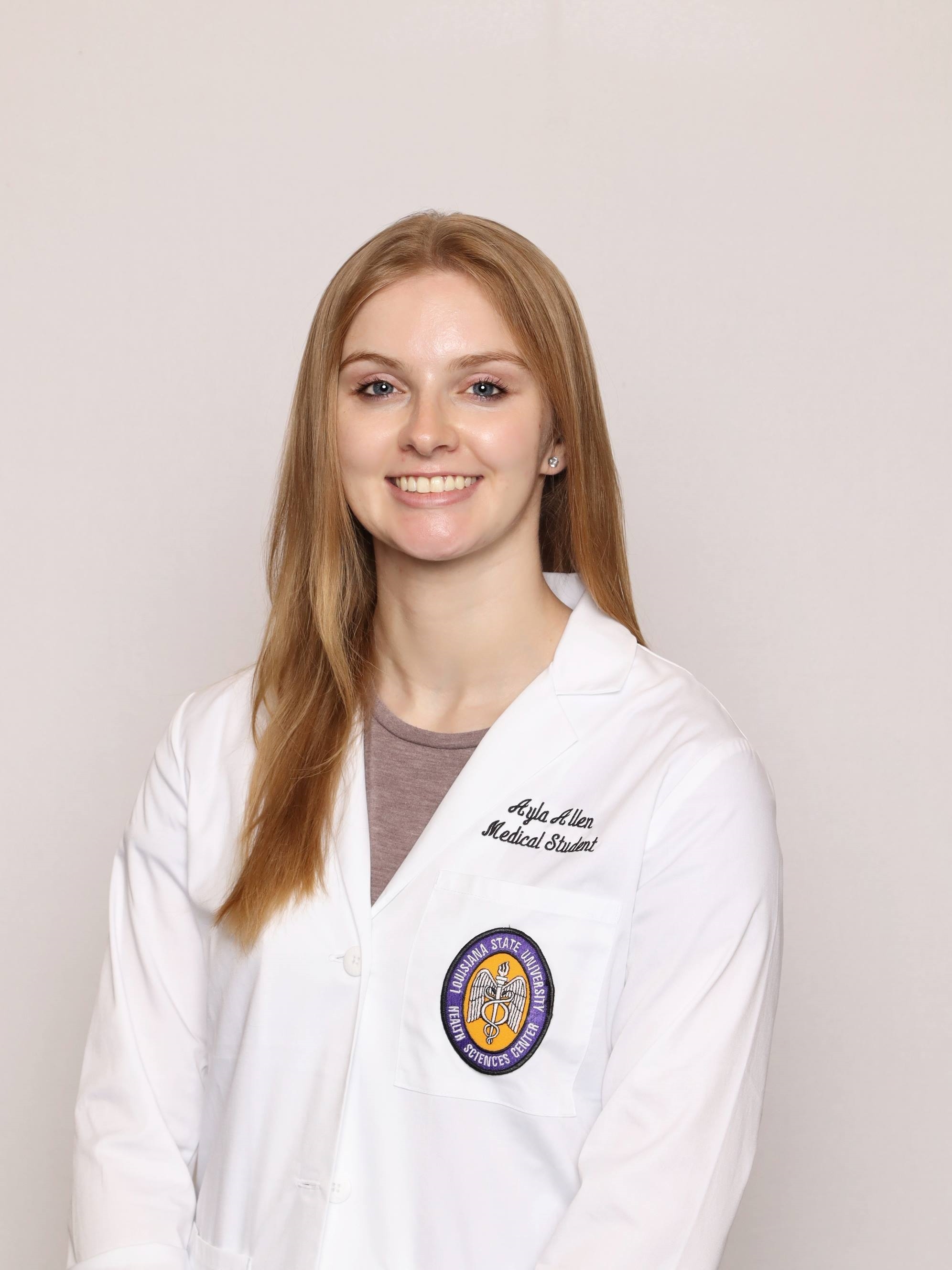 Professional medical school headshot showing a person wearing a white medical coat with embroidered text identifying the wearer as a medical student and a Louisiana State University Health Sciences Center emblem on the chest pocket. The subject is wearing a light-colored top beneath the coat and small stud earrings. The portrait is set against a plain light gray background with even, professional lighting.