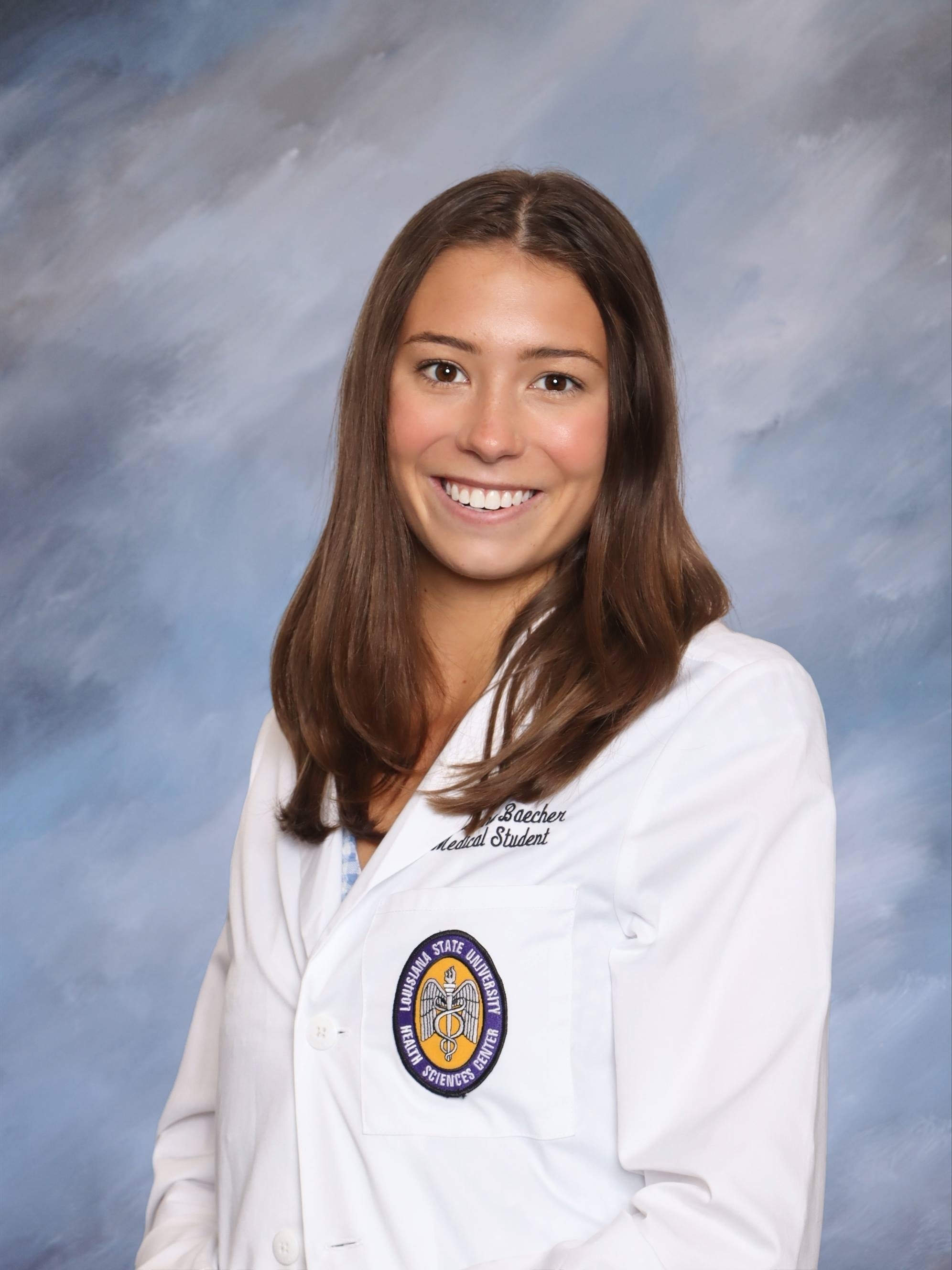 Professional medical school headshot showing a person in a white medical coat with a Louisiana State University Health Sciences Center emblem on the pocket. Visible embroidered text on the coat includes the title “Medical Student.” The image is evenly lit with a neutral blue studio backdrop that keeps the focus on the subject.