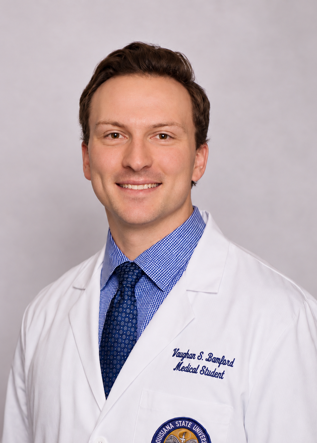 Professional medical school headshot showing a person wearing a white medical coat over a blue checkered dress shirt and blue patterned tie. Visible embroidered text on the coat includes the title “Medical Student,” and a Louisiana State University Health Sciences Center emblem is displayed on the pocket. The subject is facing the camera against a plain light gray studio background with even lighting.