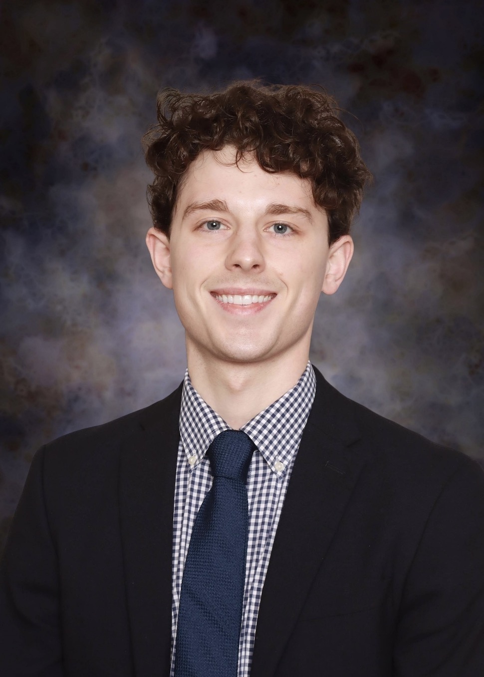 Professional headshot showing a person wearing a dark suit jacket, checkered dress shirt, and dark blue tie. The subject is facing the camera against a dark blue and gray mottled studio background. The image is evenly lit and cropped from the chest up in a formal studio portrait style.