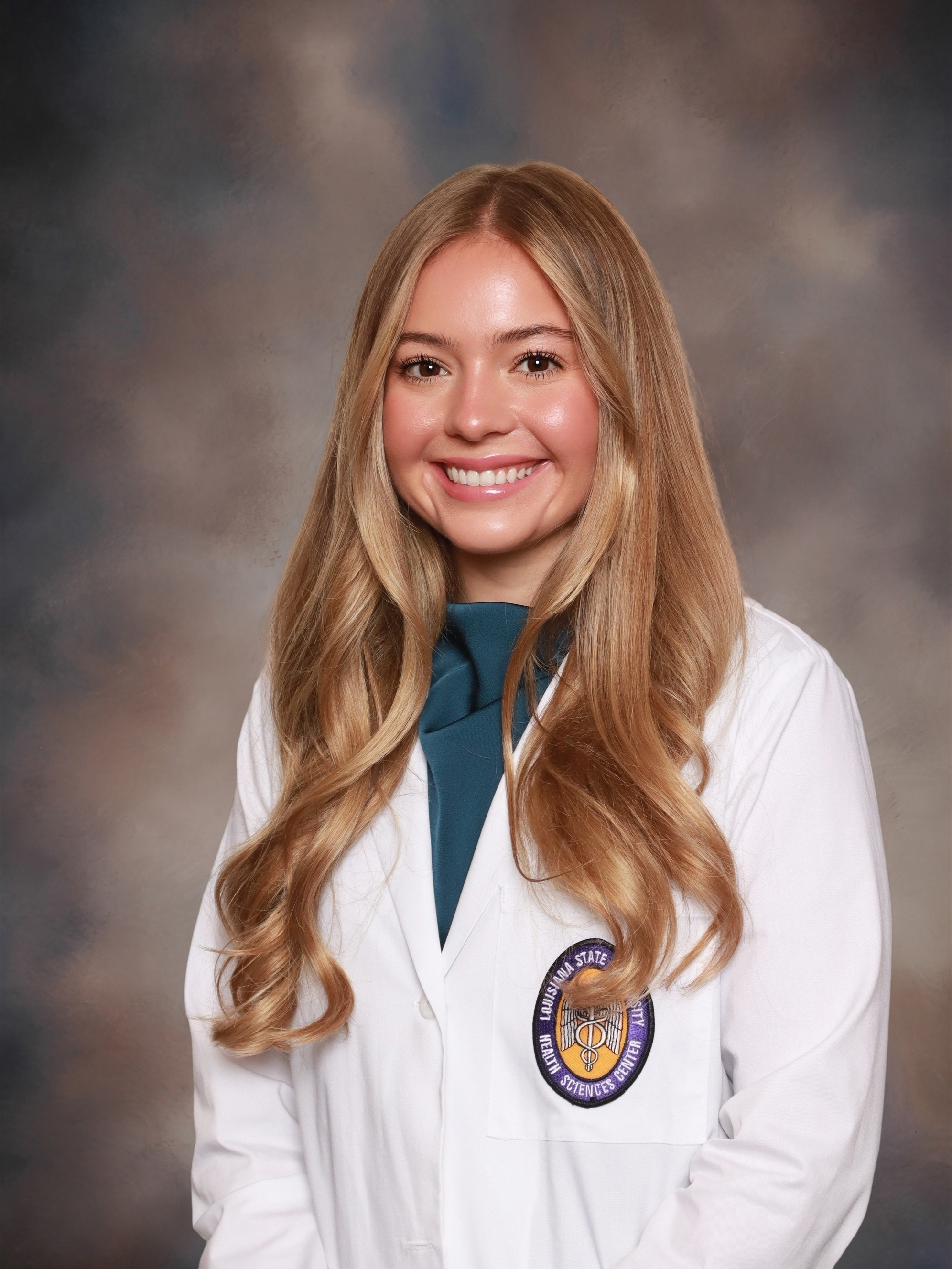 Professional medical school headshot showing a person wearing a white medical coat with a Louisiana State University Health Sciences Center emblem on the chest pocket. The subject is wearing a teal high-neck top beneath the coat. The portrait is set against a softly blended gray and brown studio background with even, professional lighting.