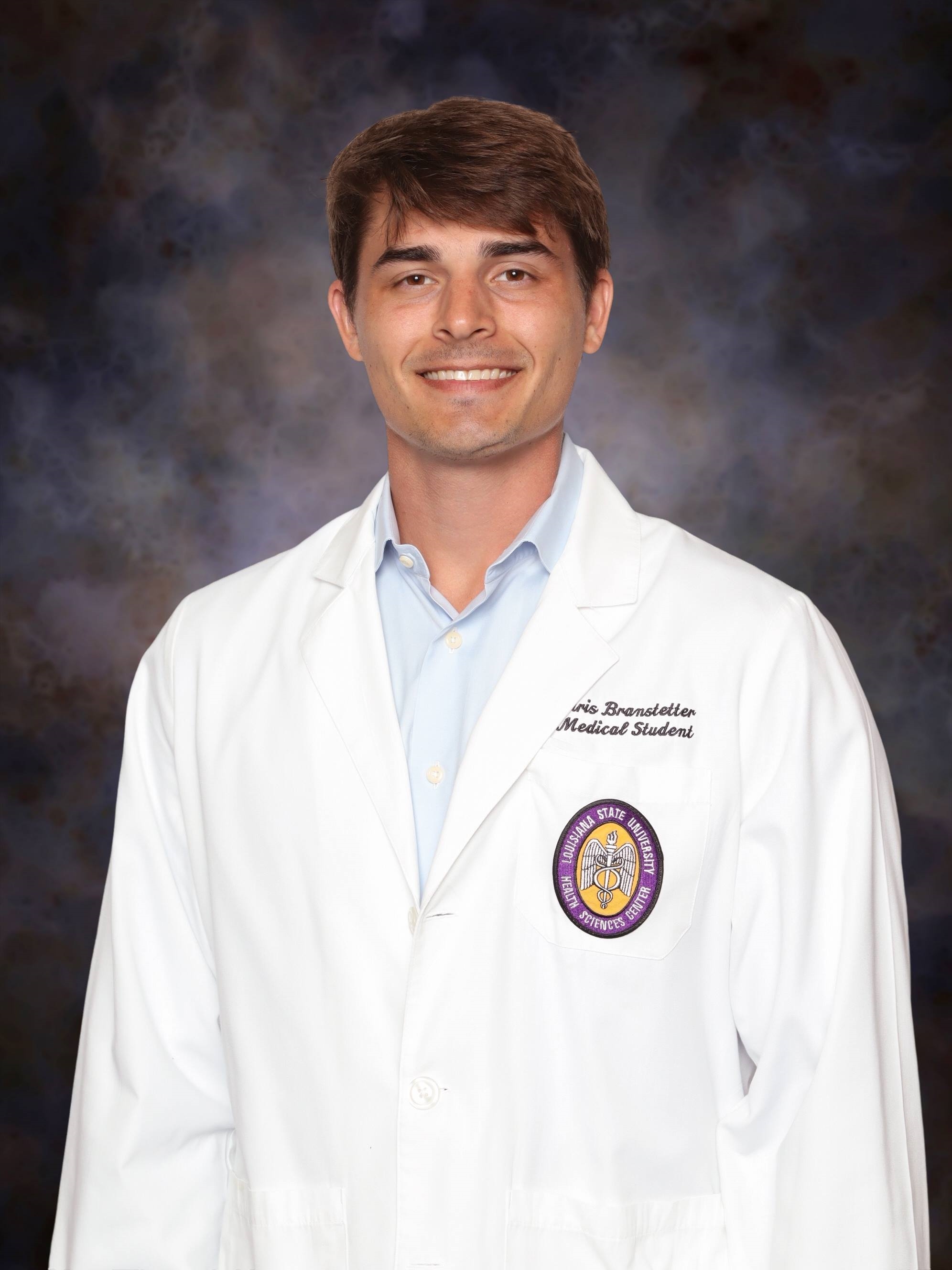 Professional medical school headshot showing a person wearing a white medical coat over a light blue button-down shirt. Visible embroidered text on the coat includes the title “Medical Student,” and a Louisiana State University Health Sciences Center emblem is displayed on the chest pocket. The subject is facing the camera against a dark blue and gray mottled studio background with even, professional lighting.