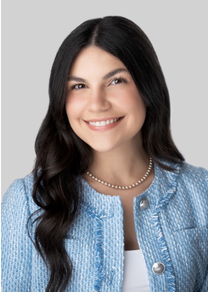 Professional headshot of a person against a light gray studio background. The person has long dark wavy hair and is wearing a light blue textured jacket over a white top, along with a short pearl necklace. The portrait is framed from the chest up, with the person facing the camera directly. Soft, even lighting creates clear visibility of facial features and clothing details, while the plain background keeps the focus on the subject.