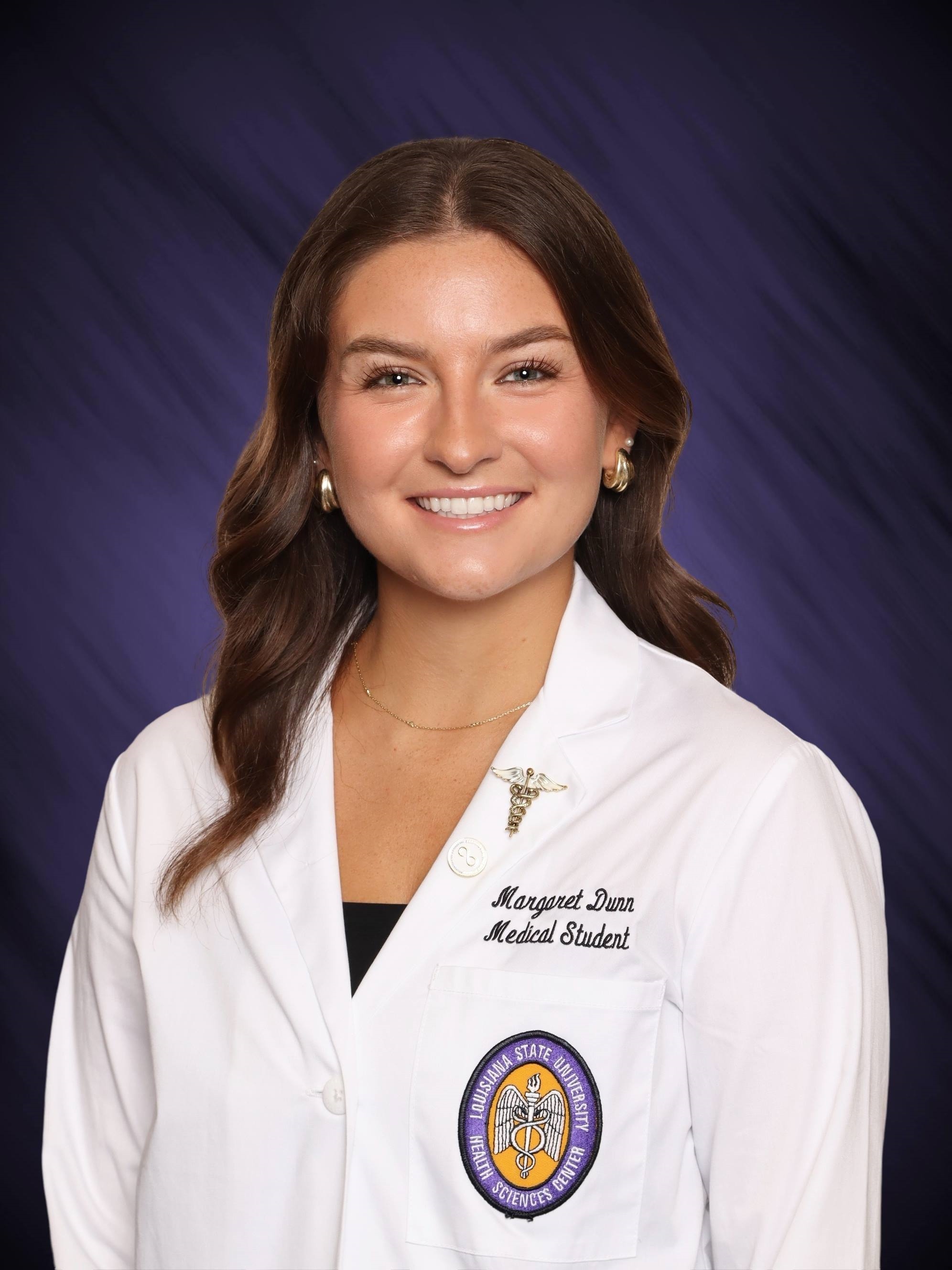 Professional medical school headshot showing a person wearing a white medical coat over a dark top. Visible embroidered text on the coat includes the title “Medical Student,” and a Louisiana State University Health Sciences Center emblem is displayed on the chest pocket. Two lapel pins, including a caduceus pin and a circular pin, are attached to the coat. The subject is wearing earrings and a delicate necklace. The portrait is set against a dark blue studio background with subtle diagonal texture and even, professional lighting.
