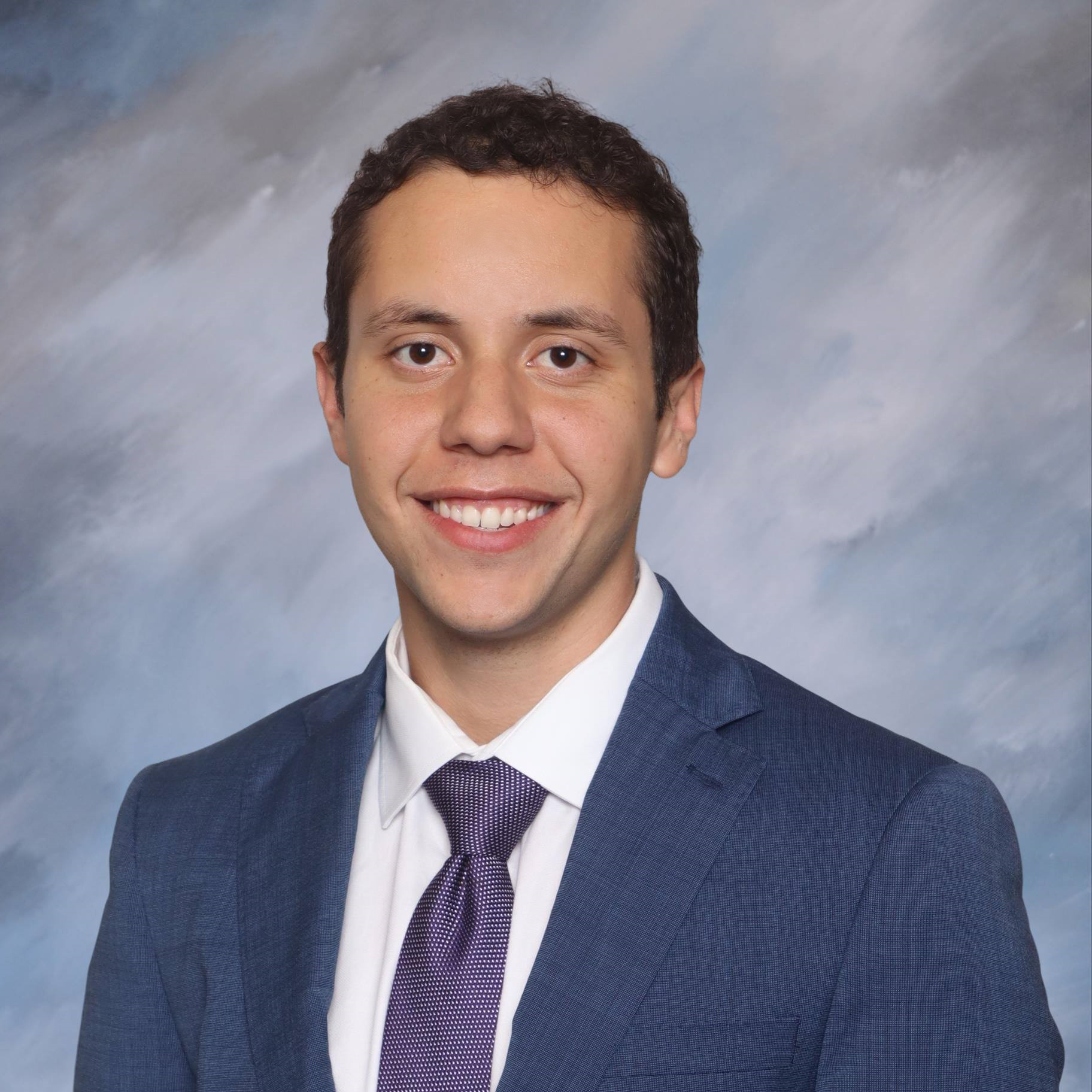 Professional headshot showing a person wearing a blue suit jacket, white dress shirt, and purple tie. The subject is facing the camera against a softly textured light blue and gray studio background. The image is evenly lit and tightly framed around the head and shoulders.