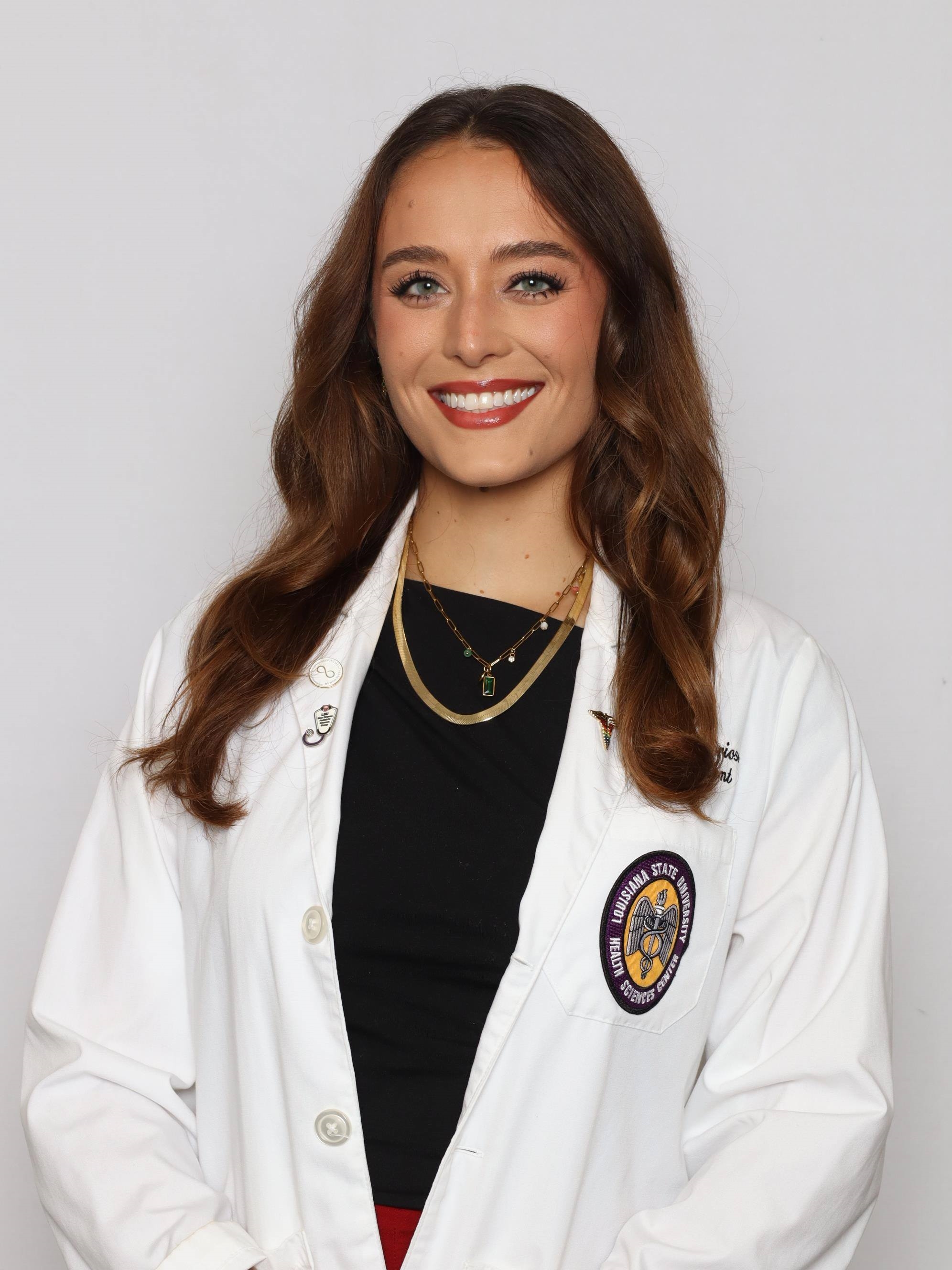 Professional medical school headshot showing a person wearing a white medical coat over a black top. The coat features a Louisiana State University Health Sciences Center emblem on the chest pocket and multiple lapel pins. The subject is wearing layered necklaces and is posed against a plain light gray background. The image is evenly lit and photographed in a professional studio style.