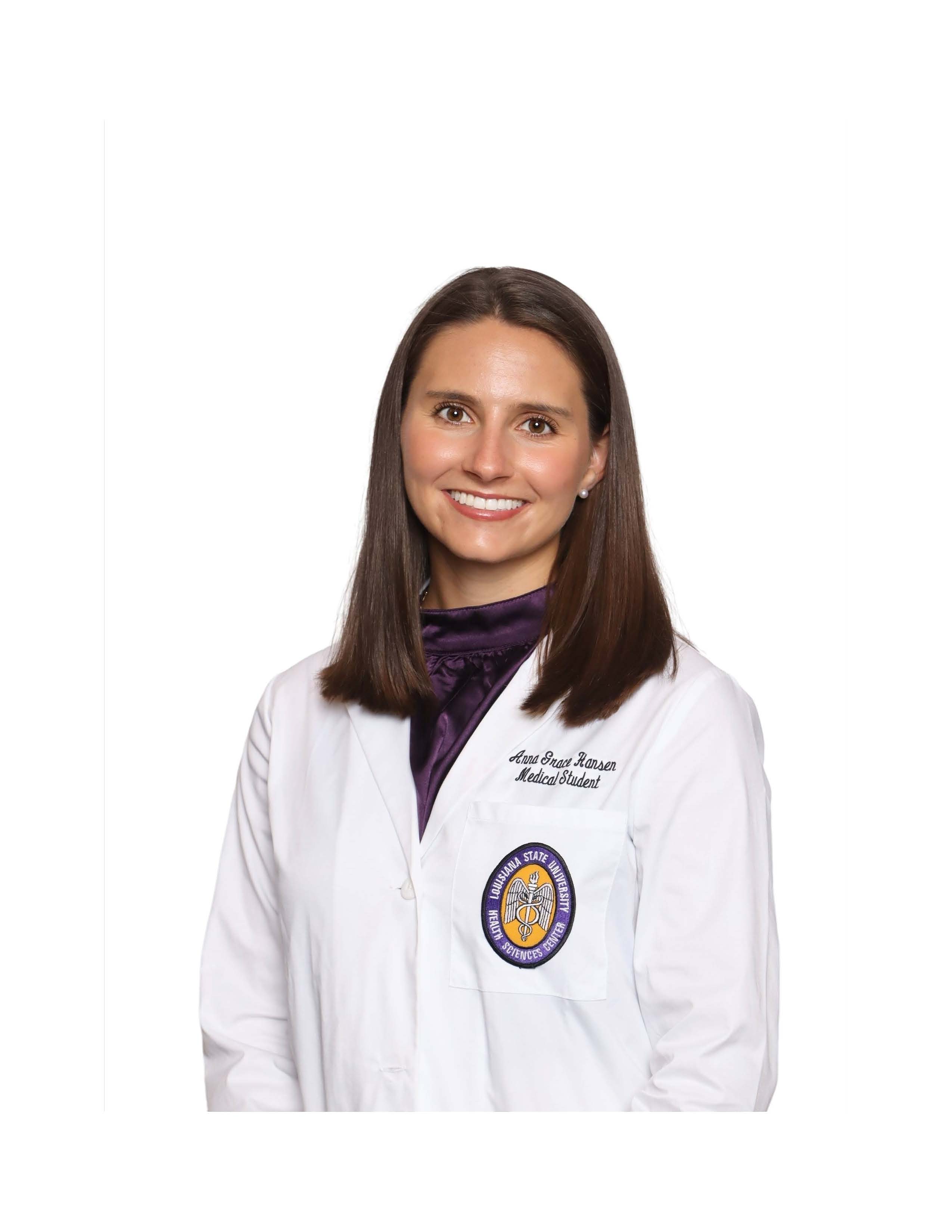 Professional headshot of a person against a light gray studio background. The person is wearing a white medical student coat over a dark purple high-neck blouse. An embroidered Louisiana State University Health Sciences Center patch is visible on the left chest pocket of the coat. The portrait is framed from the waist up, with the person facing slightly toward the camera. The image is evenly lit, and the plain background provides strong contrast that keeps attention on the subject and clothing details.