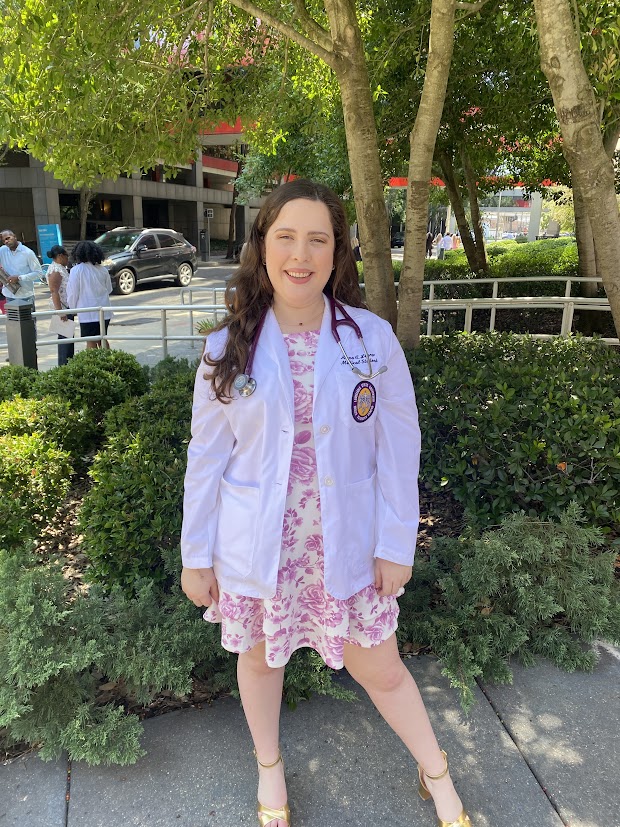 Professional medical school portrait showing a person wearing a white medical coat with a Louisiana State University Health Sciences Center emblem on the chest pocket and a stethoscope around the neck. The subject is standing outdoors on a sidewalk in front of landscaped greenery and trees, wearing a pink floral dress beneath the coat. Buildings, parked vehicles, and several people are visible in the background. The image is lit by natural daylight.