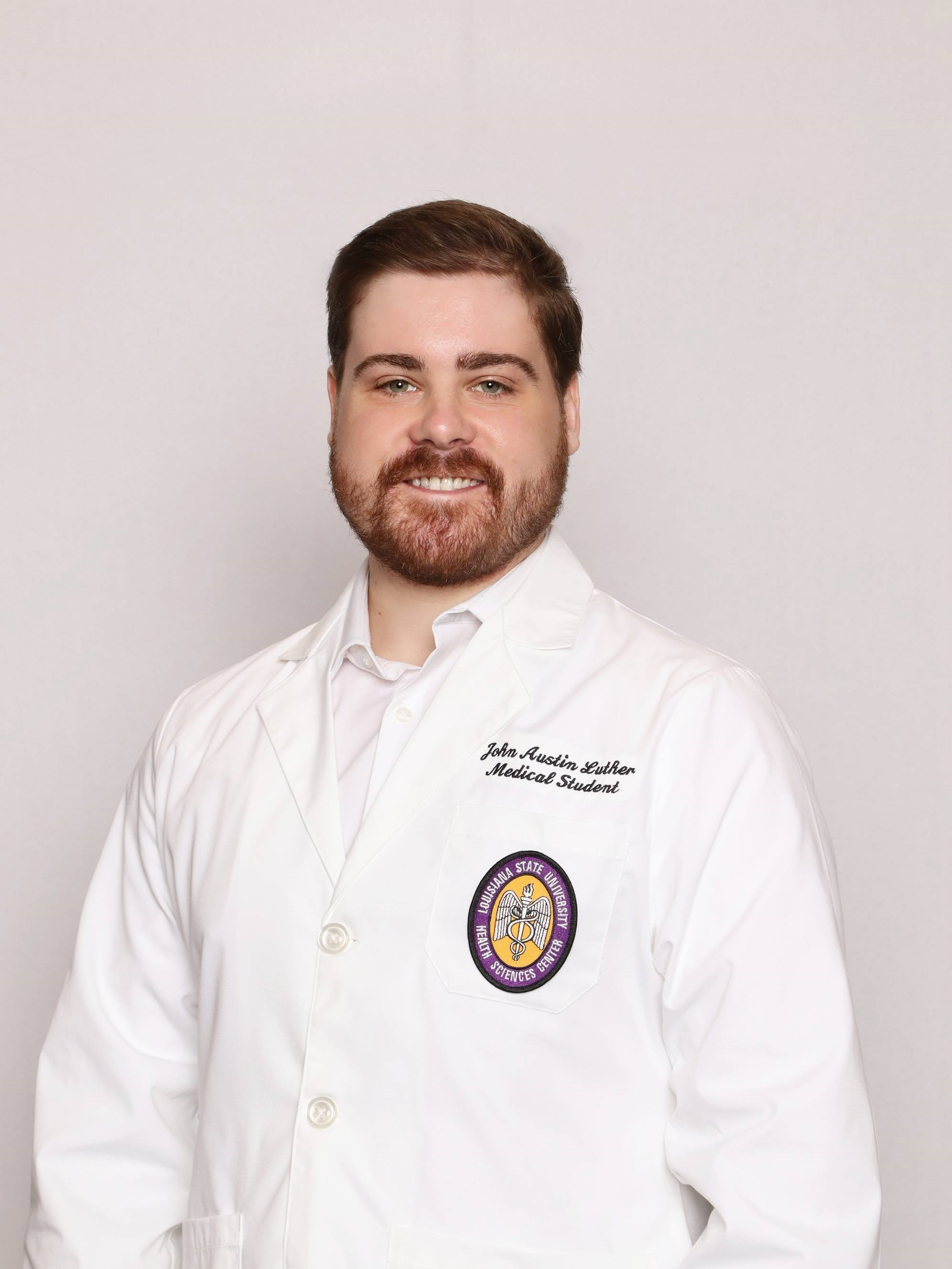 Professional medical school portrait showing a person wearing a white medical coat over a white collared shirt. Visible embroidered text on the coat includes the title “Medical Student,” and a Louisiana State University Health Sciences Center emblem is displayed on the chest pocket. The subject has a beard and mustache and is facing the camera against a plain light gray background. The image is evenly lit and presented in a formal studio portrait style.