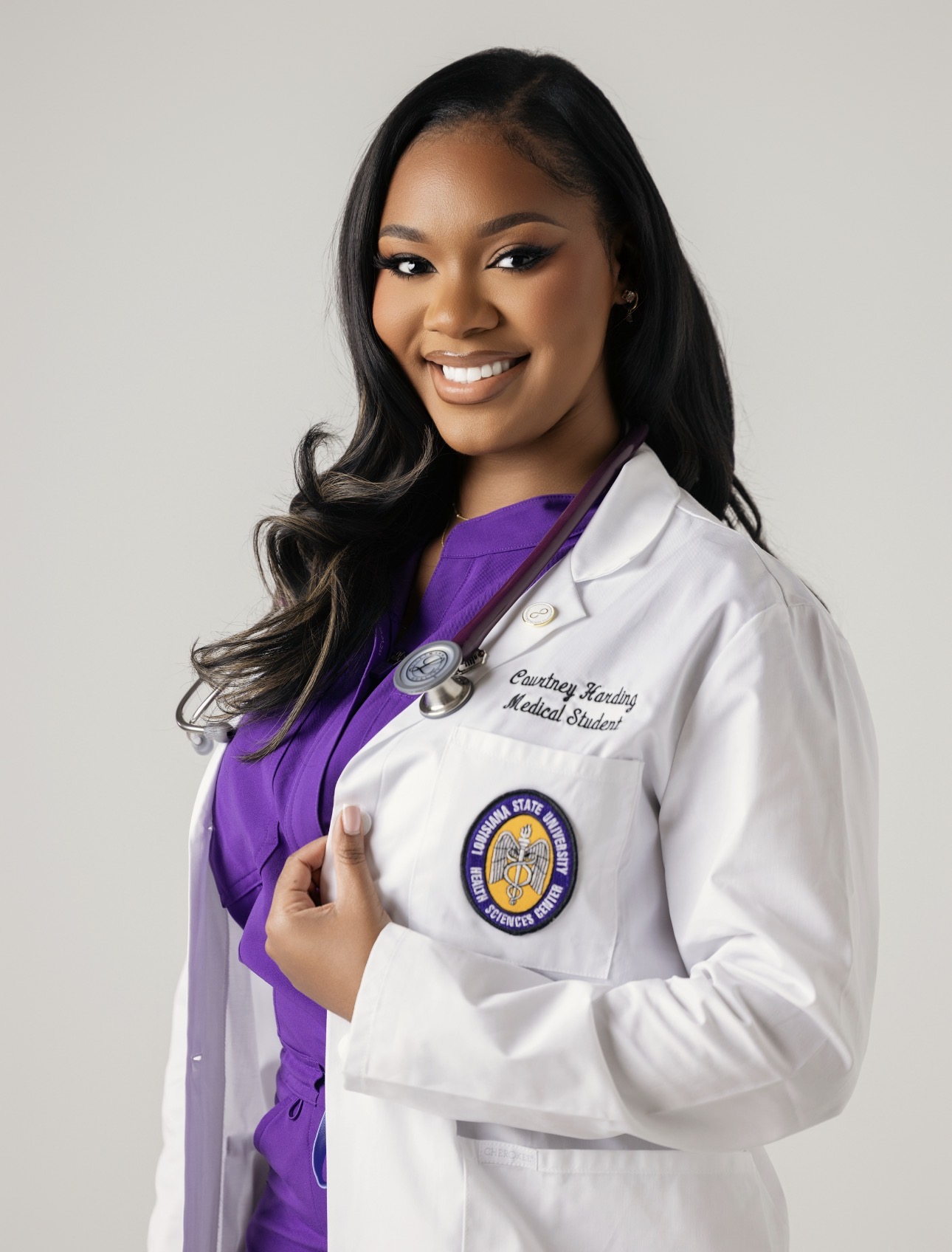 Professional medical school portrait of a person wearing a white medical coat over purple scrubs with a stethoscope around the neck, against a light gray background.