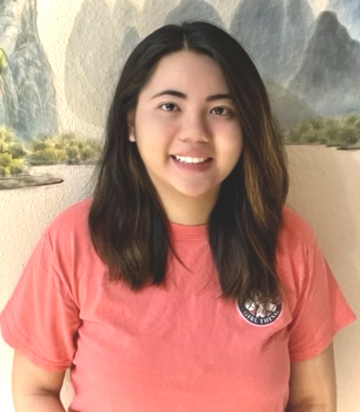 Casual portrait of a person wearing a coral-pink T-shirt, standing indoors in front of a mural depicting mountains and a landscape.
