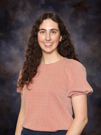 Professional studio portrait of a person wearing a patterned light pink blouse against a dark blue-gray studio background.