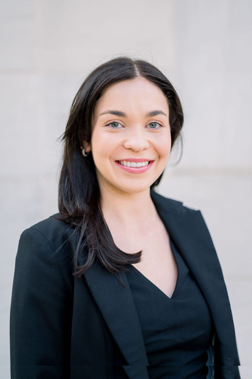 Professional outdoor-style portrait of a person wearing a black blazer and black top against a light stone background.