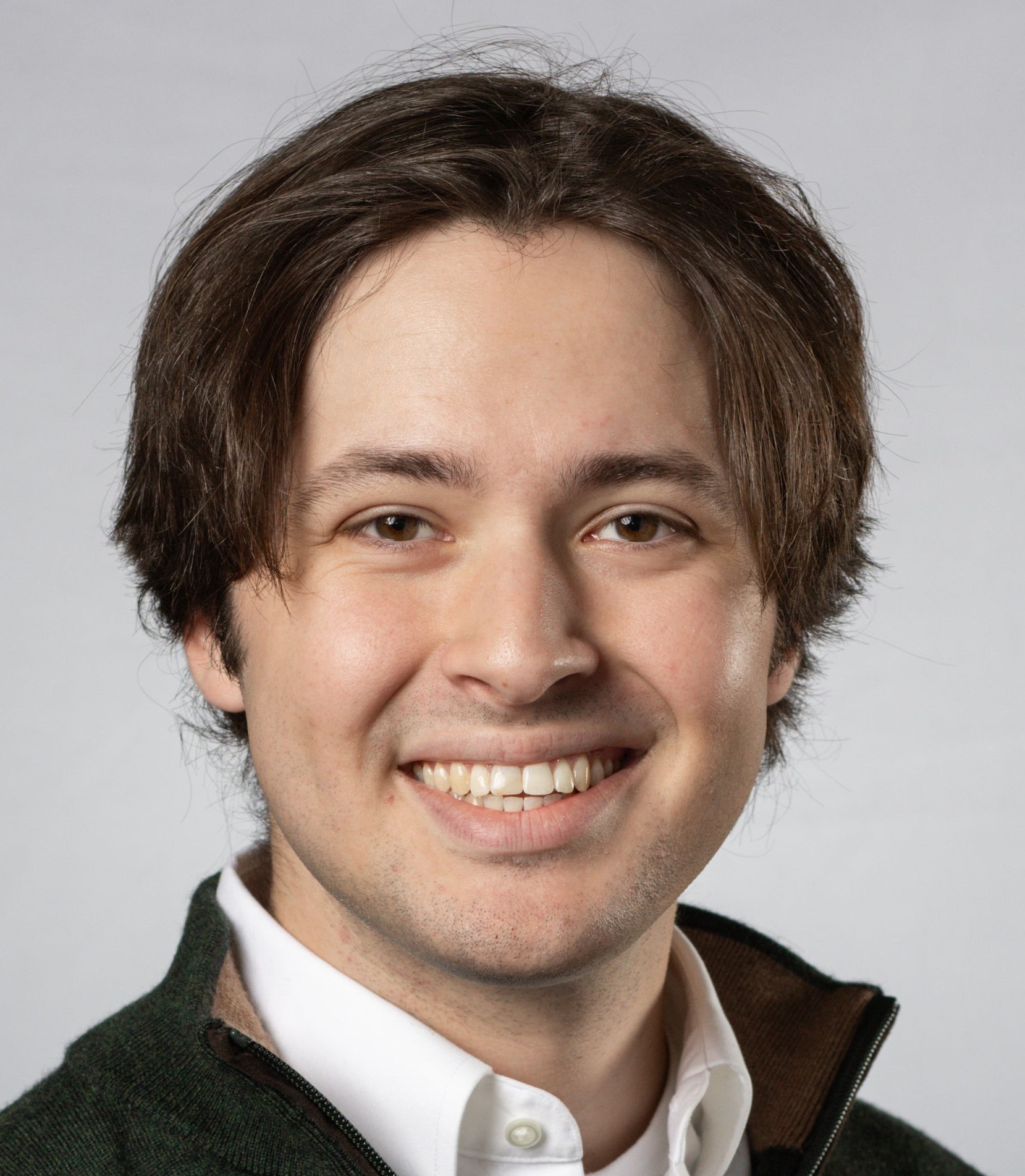 Professional headshot of a person wearing a white collared shirt and a dark quarter-zip sweater against a light gray background.