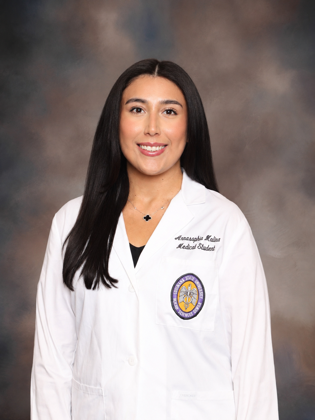 Professional medical school headshot showing a person wearing a white medical coat with embroidered text identifying the wearer as a medical student and a Louisiana State University Health Sciences Center emblem on the chest pocket. The subject is wearing a dark top and a necklace and is facing the camera against a softly blended gray and brown studio background. The image is evenly lit with a clear focus on the subject.