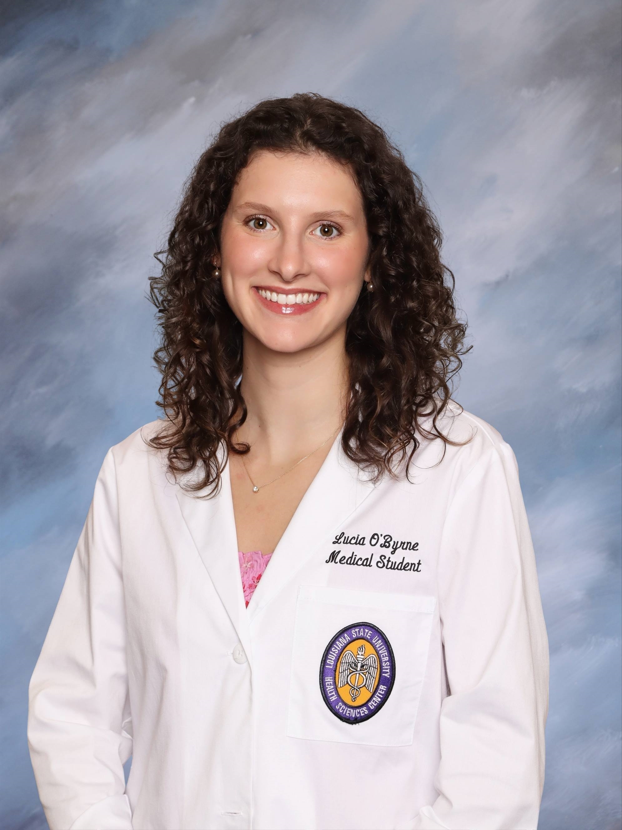 Professional medical school headshot showing a person wearing a white medical coat with embroidered text identifying the wearer as a medical student and a Louisiana State University Health Sciences Center emblem on the chest pocket. The subject is wearing a pink top beneath the coat and a small necklace. The portrait is set against a softly textured light blue studio background with even, professional lighting.