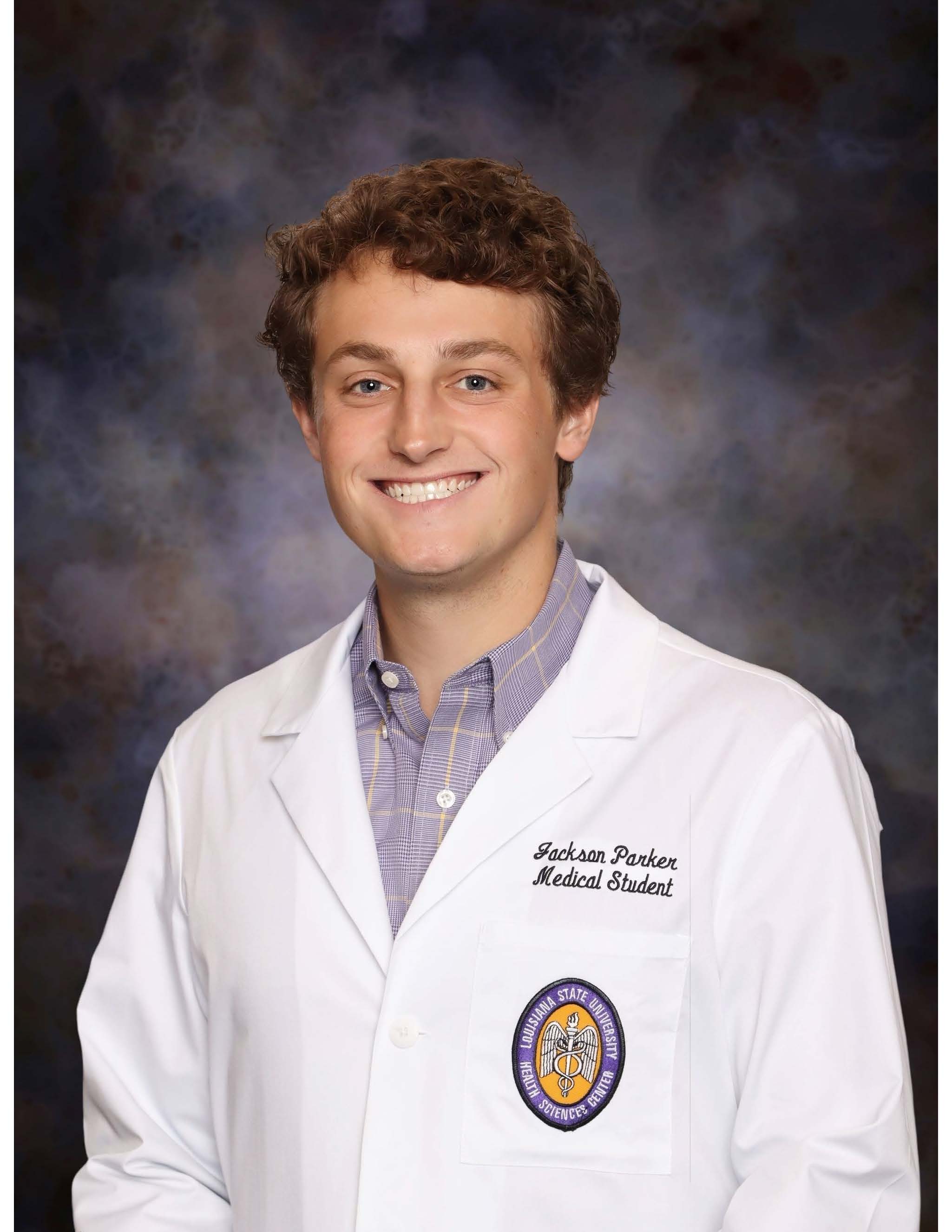Professional medical school headshot showing a person wearing a white medical coat over a light-colored checkered button-down shirt. Visible embroidered text on the coat includes the title “Medical Student,” and a Louisiana State University Health Sciences Center emblem is displayed on the chest pocket. The subject is facing the camera against a dark blue and gray mottled studio background with even, professional lighting.