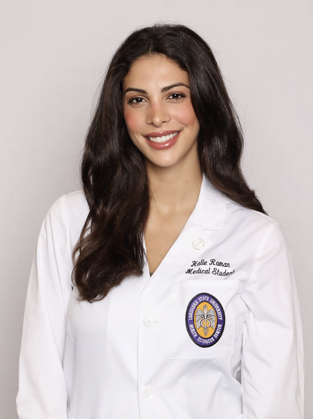 Professional medical school headshot showing a person wearing a white medical coat with embroidered text identifying the wearer as a medical student and a Louisiana State University Health Sciences Center emblem on the chest pocket. A lapel pin is attached to the coat. The subject is facing the camera against a plain light gray background with even, professional lighting.