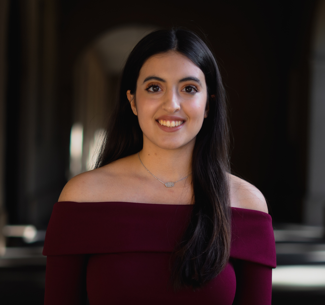 Professional portrait of a person positioned in the center of the frame against a softly blurred indoor architectural background with dark archways and columns. The person has long dark hair and is wearing a burgundy off-the-shoulder top and a thin necklace with a small pendant. The image is framed from the chest up, with the person facing the camera directly. Soft natural lighting highlights the subject while the background remains out of focus, creating a clear separation between the subject and the surroundings.