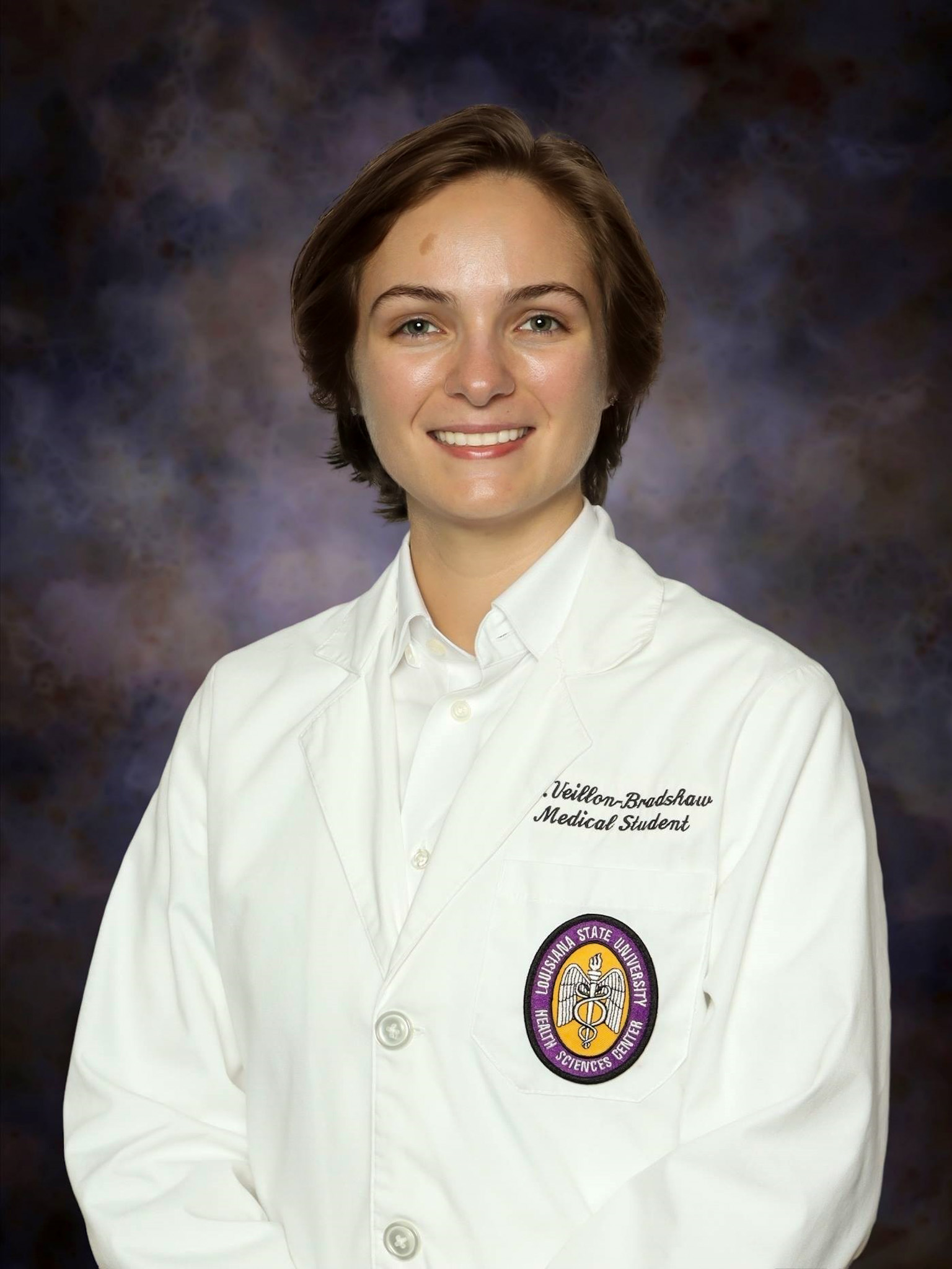 Professional medical school headshot showing a person wearing a white medical coat over a white button-down shirt. Visible embroidered text on the coat includes the title “Medical Student,” and a Louisiana State University Health Sciences Center emblem is displayed on the chest pocket. The subject is facing the camera against a dark blue and gray mottled studio background with even, professional lighting.