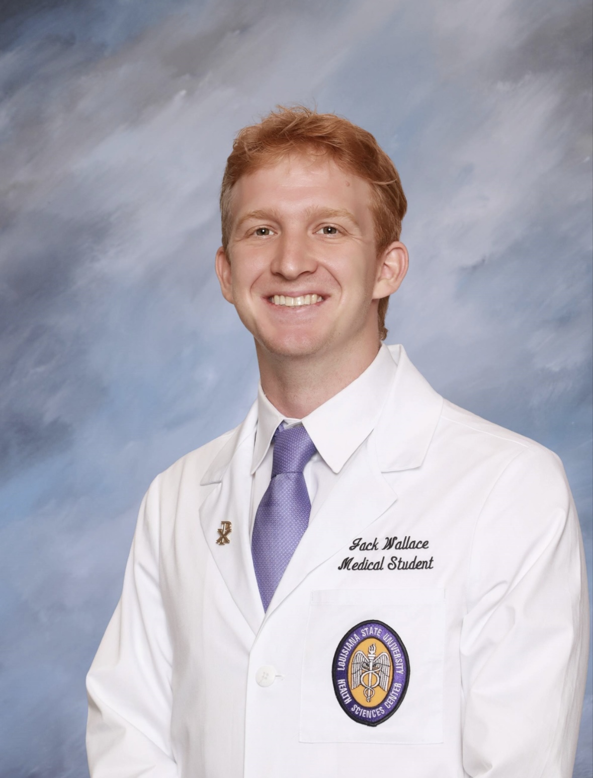 Professional medical school portrait showing a person wearing a white medical coat over a white dress shirt and a light purple tie. Visible embroidered text on the coat includes the title “Medical Student,” and a Louisiana State University Health Sciences Center emblem is displayed on the chest pocket. A small lapel pin is attached to the coat. The subject is facing the camera against a softly textured light blue and gray studio background. The image is evenly lit and presented in a formal portrait style.