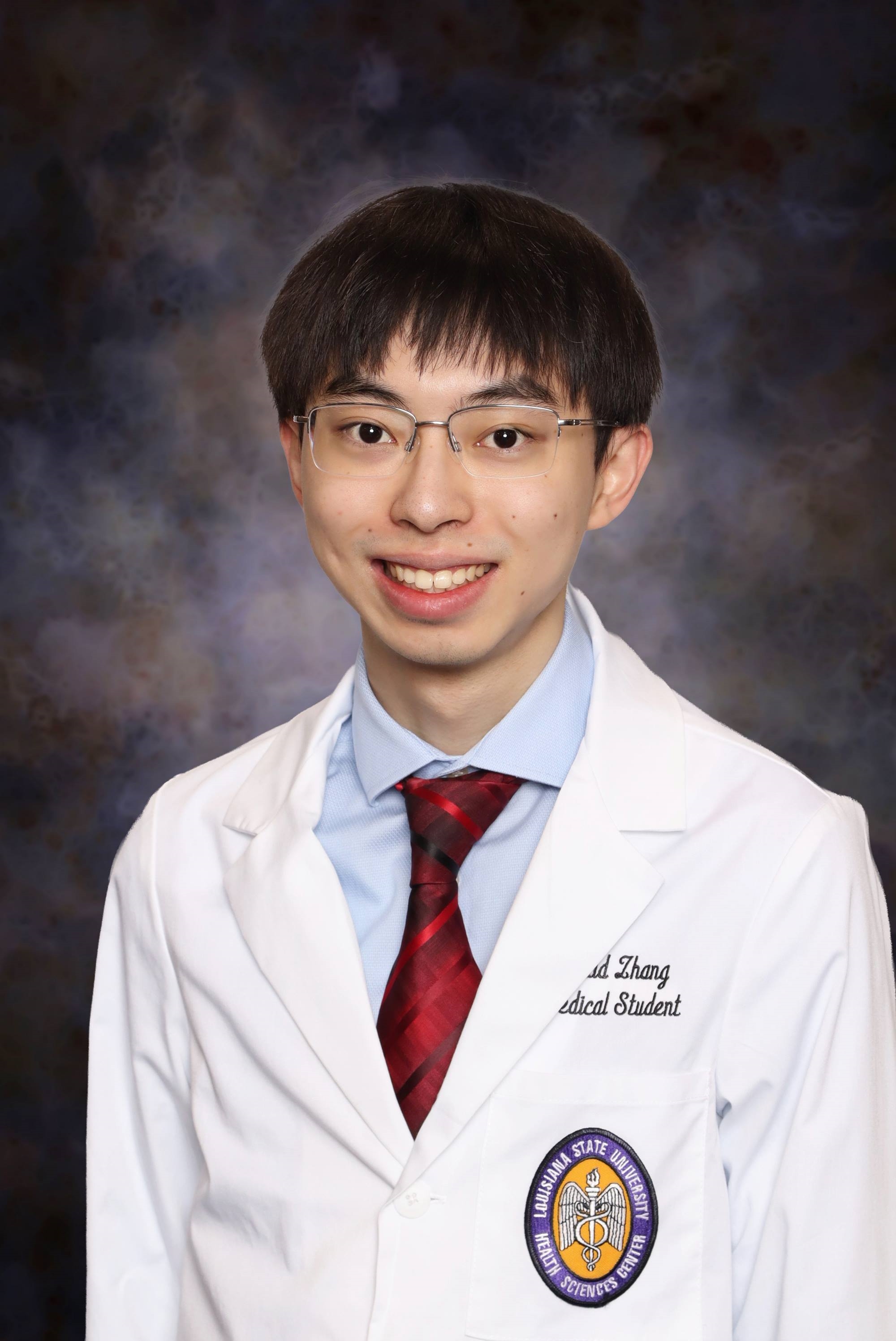 Professional medical school headshot showing a person wearing a white medical coat over a light blue dress shirt and a red patterned tie. Visible embroidered text on the coat includes the title “Medical Student,” and a Louisiana State University Health Sciences Center emblem is displayed on the chest pocket. The subject is wearing rectangular eyeglasses. The portrait is set against a dark blue and gray mottled studio background with even, professional lighting.