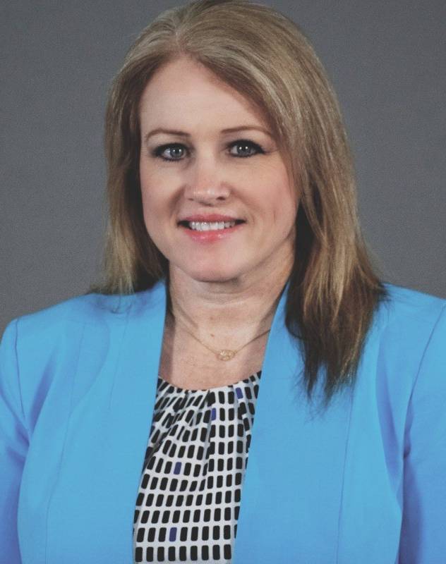 Professional headshot of a person photographed against a gray studio background. The person is wearing a light blue blazer over a patterned black-and-white blouse and a delicate necklace. The portrait is framed from the chest up, with the subject facing the camera directly. Soft studio lighting and a neutral backdrop create a formal business-profile appearance.