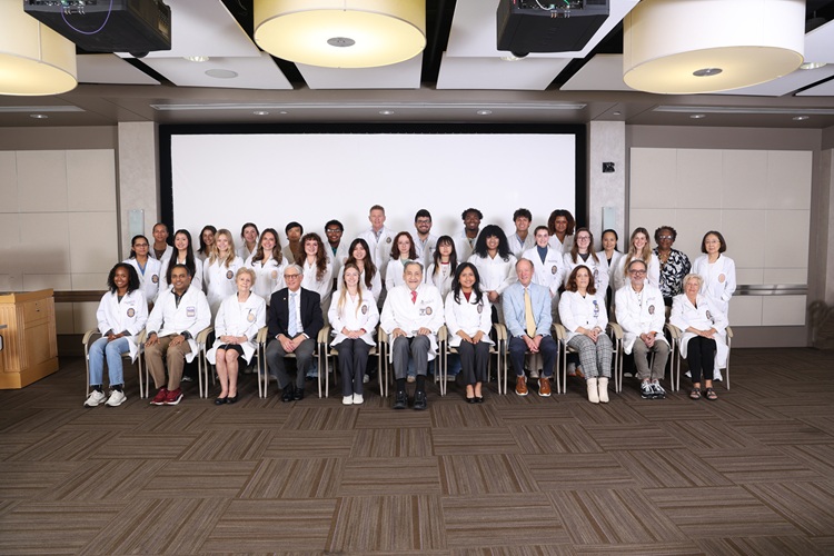 Large group portrait of approximately 35–40 people wearing white lab coats, arranged in three rows in a conference or lecture room. The front row is seated on chairs while the middle and back rows stand behind them. A large projection screen fills the center background, with modern ceiling light fixtures and mounted projectors overhead. The room has neutral-colored walls and patterned carpet flooring. The group faces the camera for a formal team or program photo.