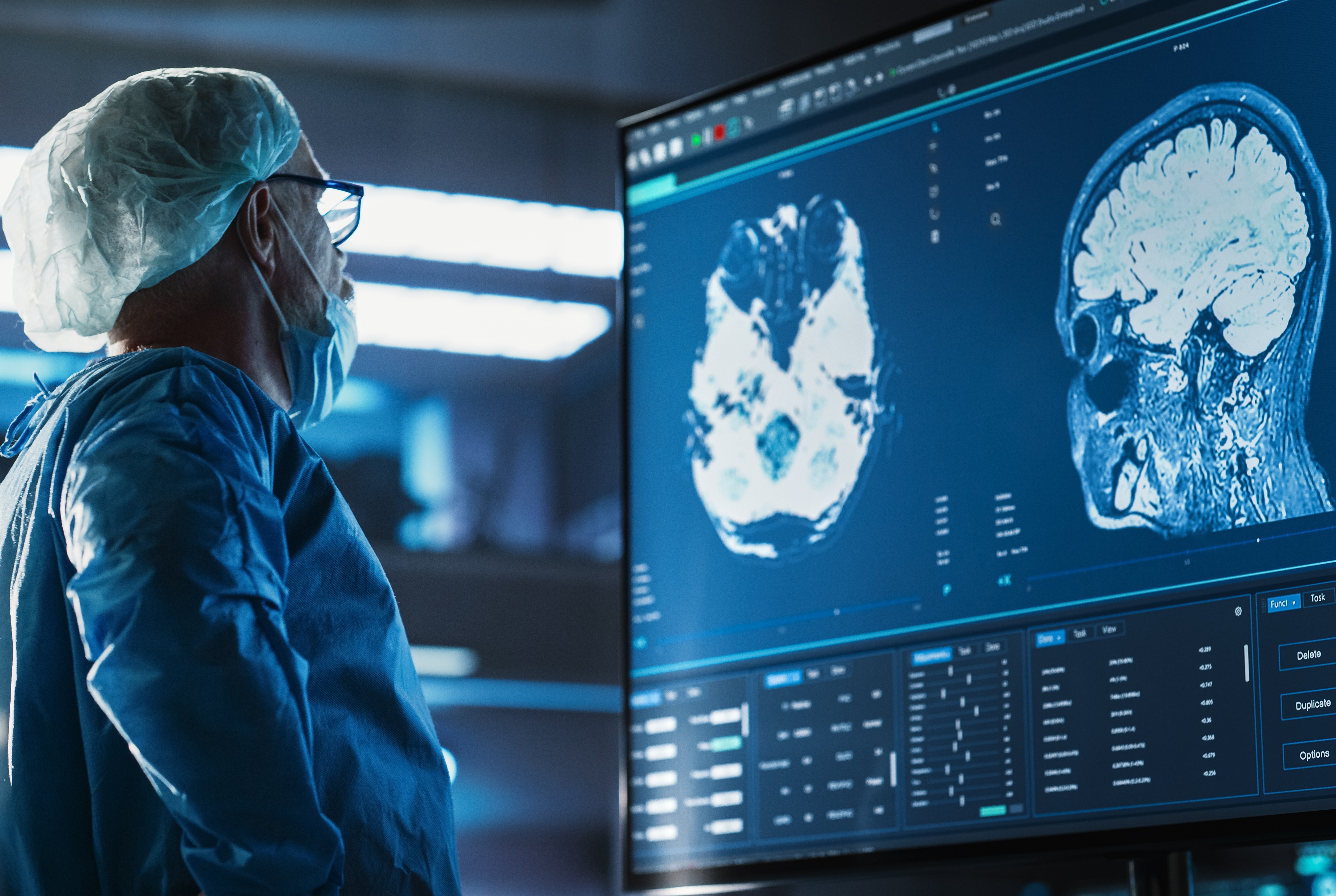 A healthcare professional wearing surgical attire, including a protective cap, mask, glasses, and clinical gown, stands in front of a large medical display showing detailed brain imaging scans. The monitor presents multiple views of brain anatomy along with diagnostic software panels and measurement data. The room is illuminated by the glow of the display and overhead clinical lighting, creating a high-tech medical environment. The image highlights the review and analysis of neuroimaging studies for diagnosis, treatment planning, or ongoing management of neurological conditions.