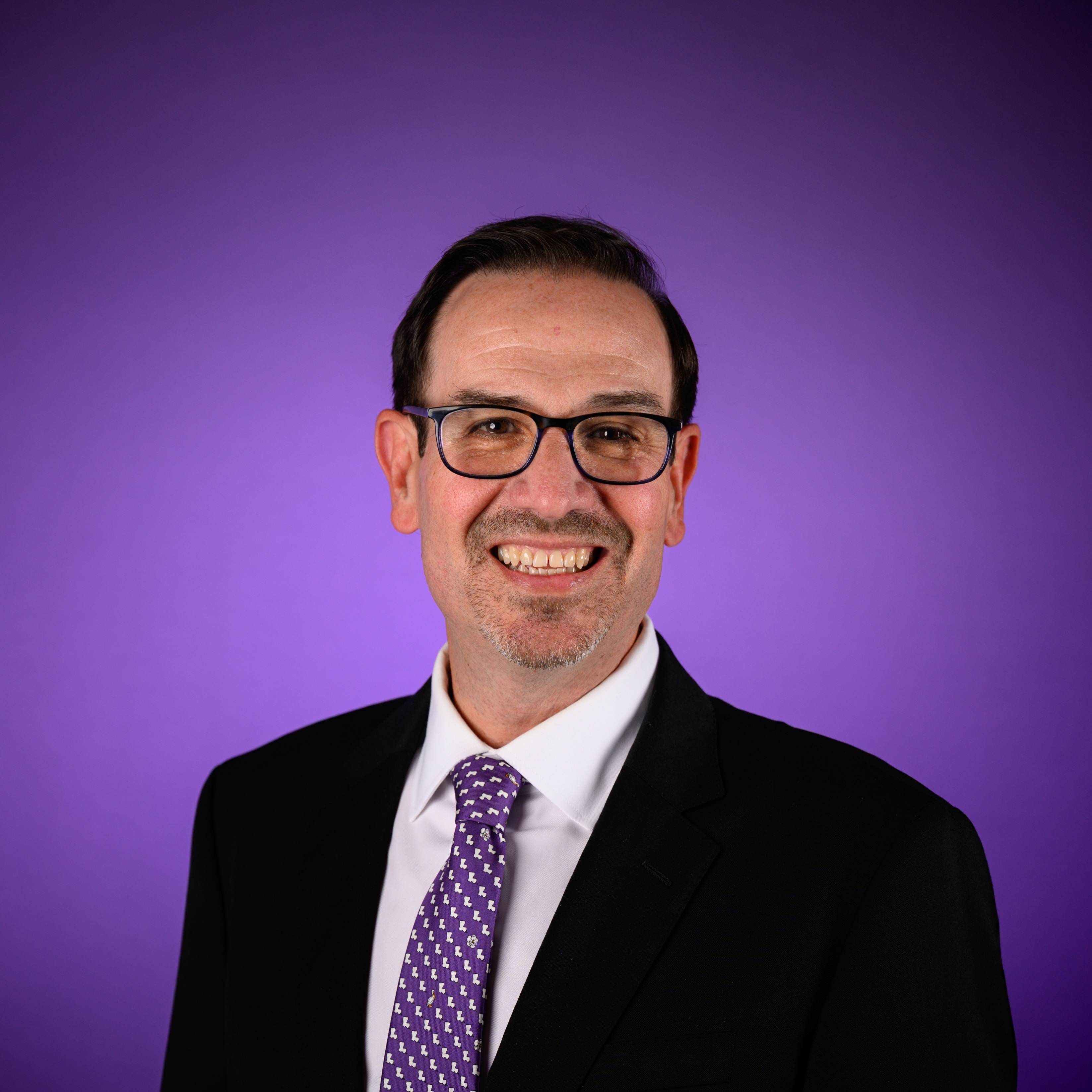 Studio portrait of a person wearing glasses, a black suit jacket, a white dress shirt, and a purple patterned tie, positioned against a smooth purple gradient background.