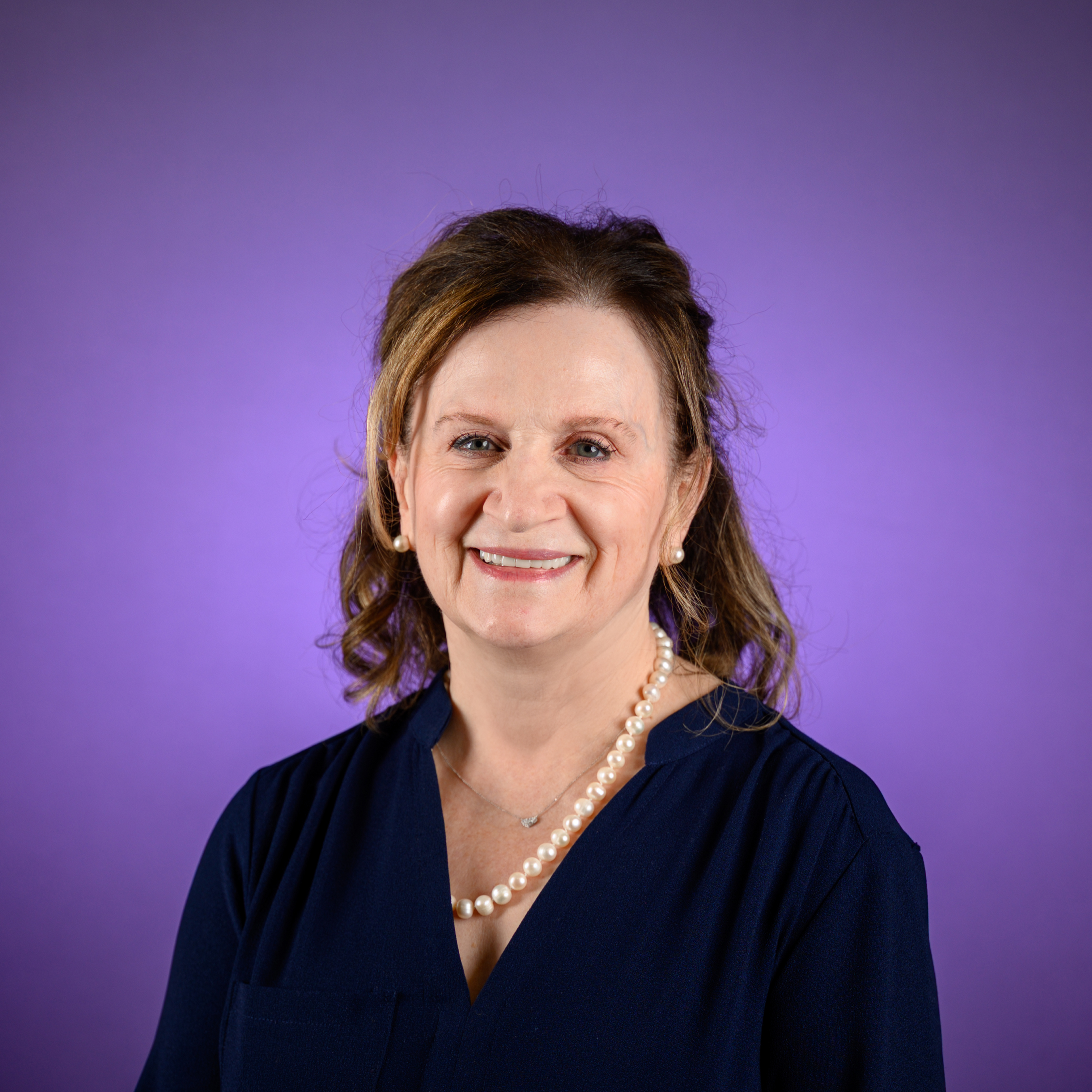 Head-and-shoulders studio portrait of an adult with medium-length hair pulled back, wearing a dark V-neck top and a single-strand pearl necklace, photographed against a solid purple background.