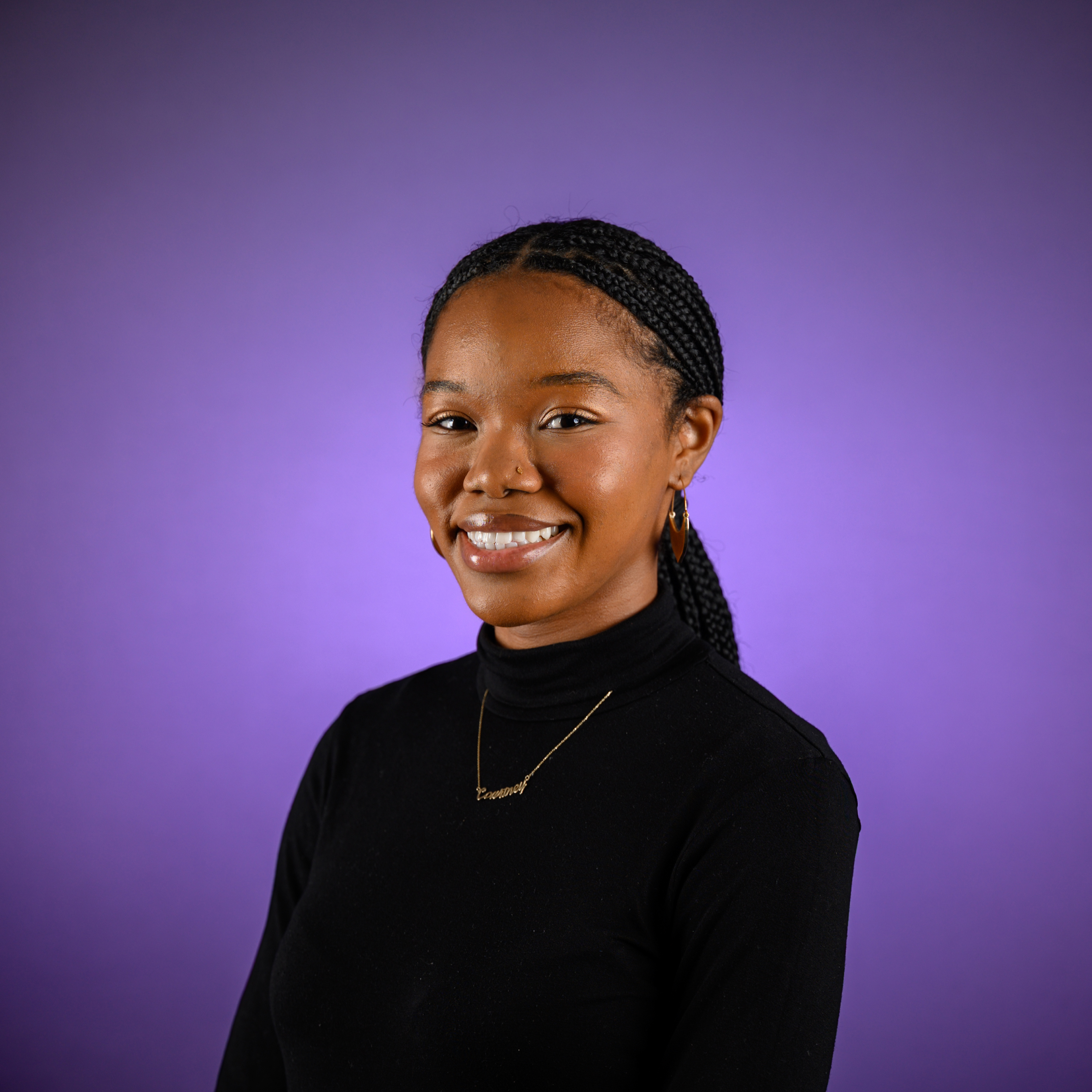 Head-and-shoulders studio portrait of an adult with braided hair pulled back, wearing a black long-sleeved top and a delicate necklace, photographed against a smooth purple background.