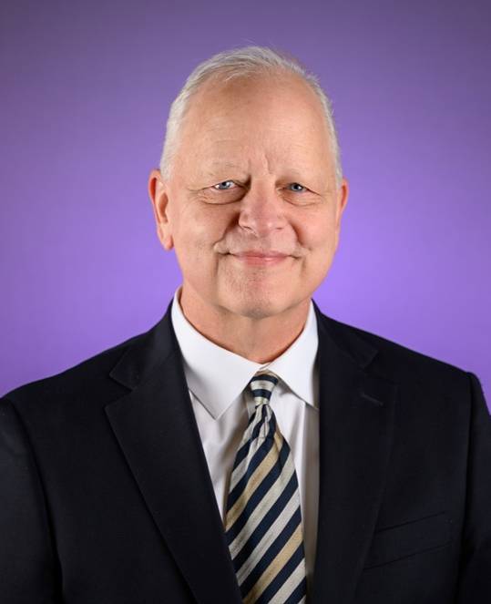 Studio portrait of a person centered against a smooth purple gradient background. The person is wearing a dark suit jacket, white dress shirt, and a diagonally striped tie. The individual has short, light-colored hair and is facing forward, looking toward the camera. The lighting is even and highlights the face and upper torso clearly against the clean, softly blended backdrop.