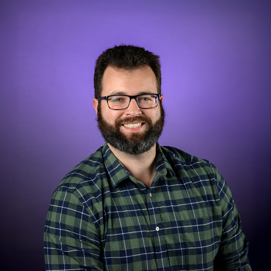 Head-and-shoulders studio portrait of an adult with short dark hair and a full beard, wearing rectangular eyeglasses and a green plaid button-up shirt, photographed against a smooth purple background.