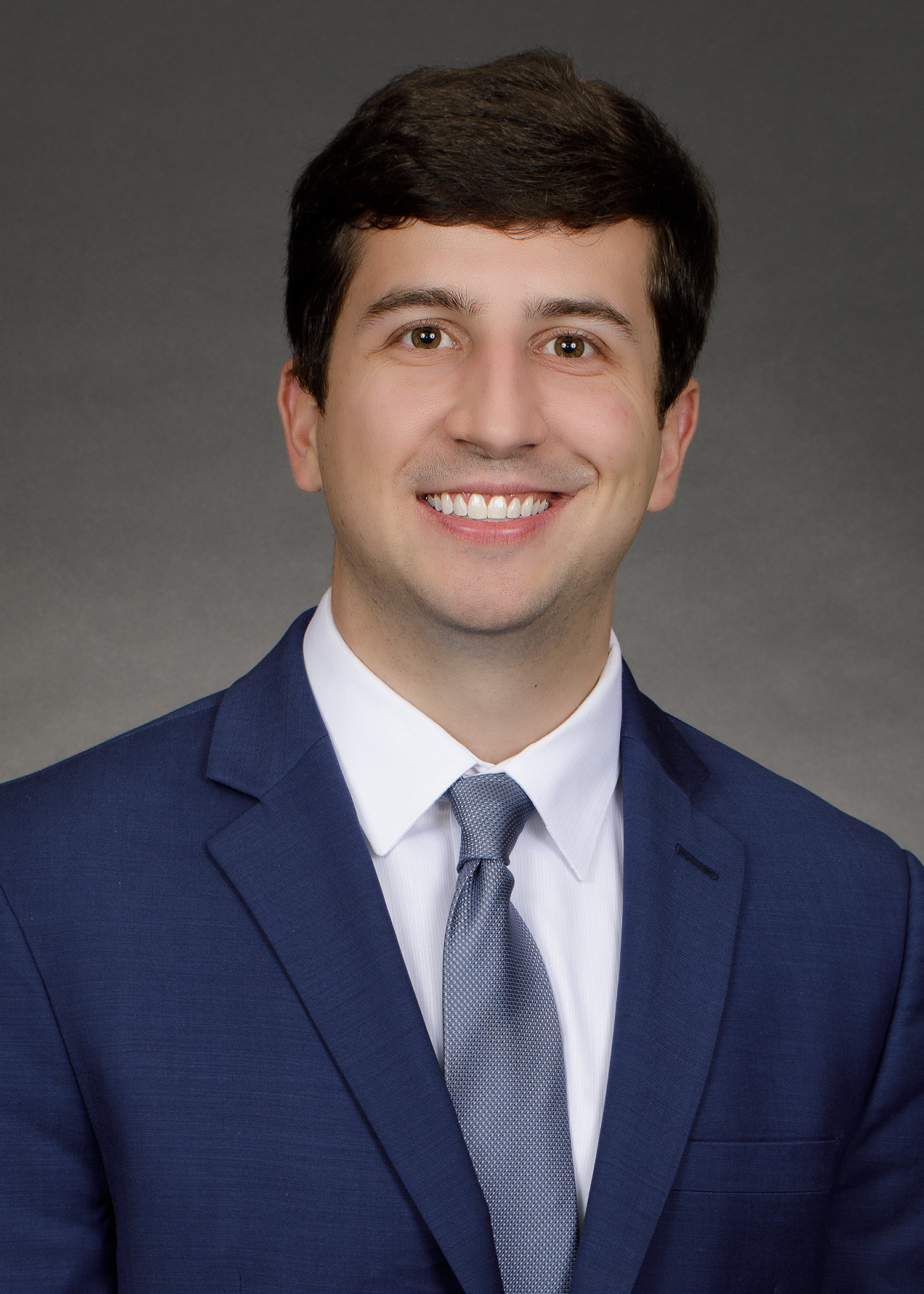 Professional head-and-shoulders portrait of a person wearing a blue suit jacket, white dress shirt, and light gray tie. The person is facing the camera and centered against a neutral gray studio background. The image is well lit, cropped from the chest upward, and resembles a formal business or professional profile photograph.