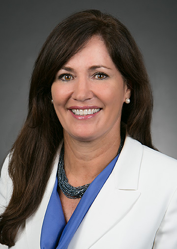 Professional head-and-shoulders portrait of an adult with long dark hair, wearing a white blazer over a blue top and a layered necklace, photographed against a neutral gray studio background.