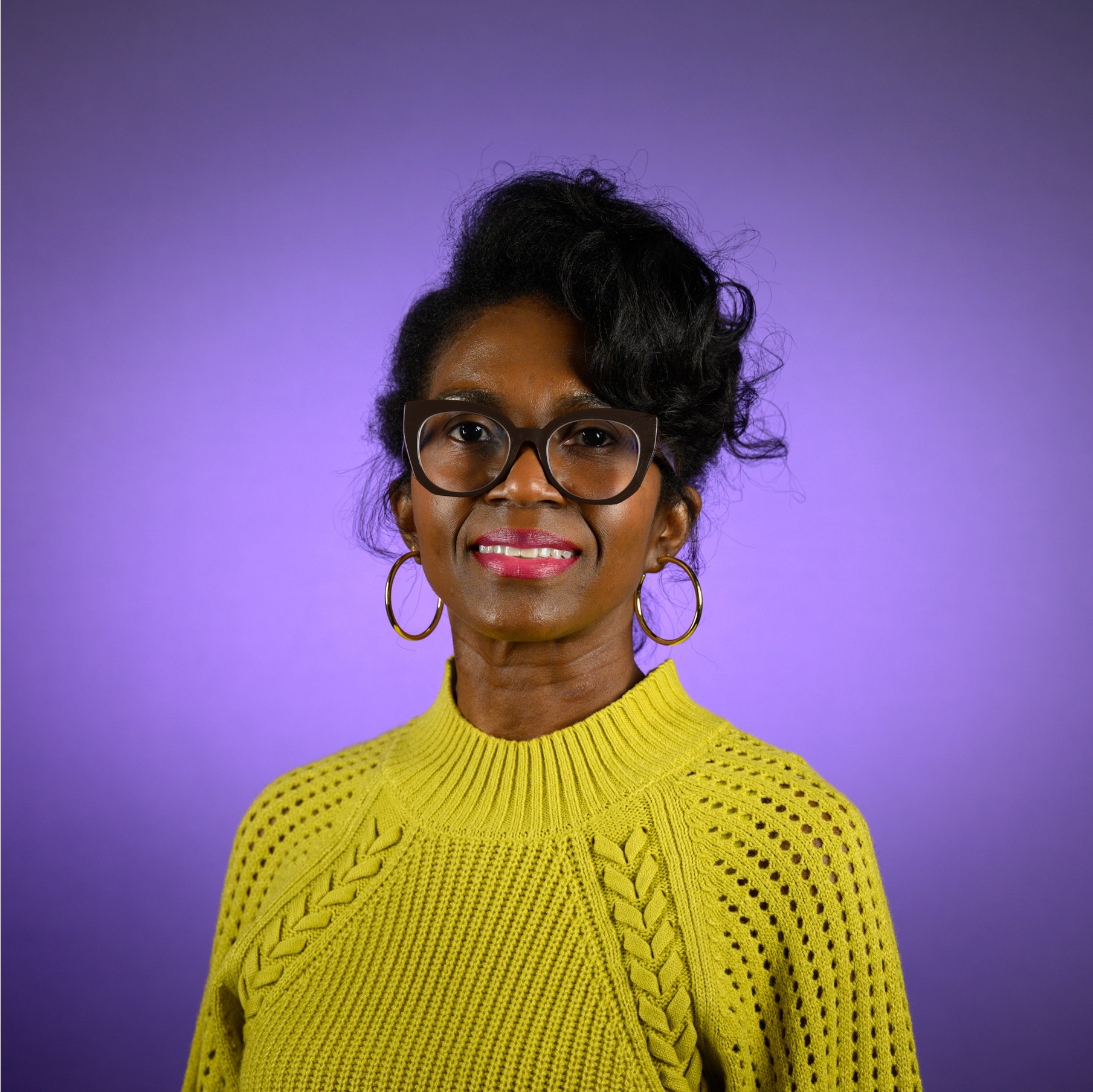 Head-and-shoulders studio portrait of an adult wearing round eyeglasses, hoop earrings, and a textured bright knit sweater, with styled hair swept upward and to the side, photographed against a smooth purple background.