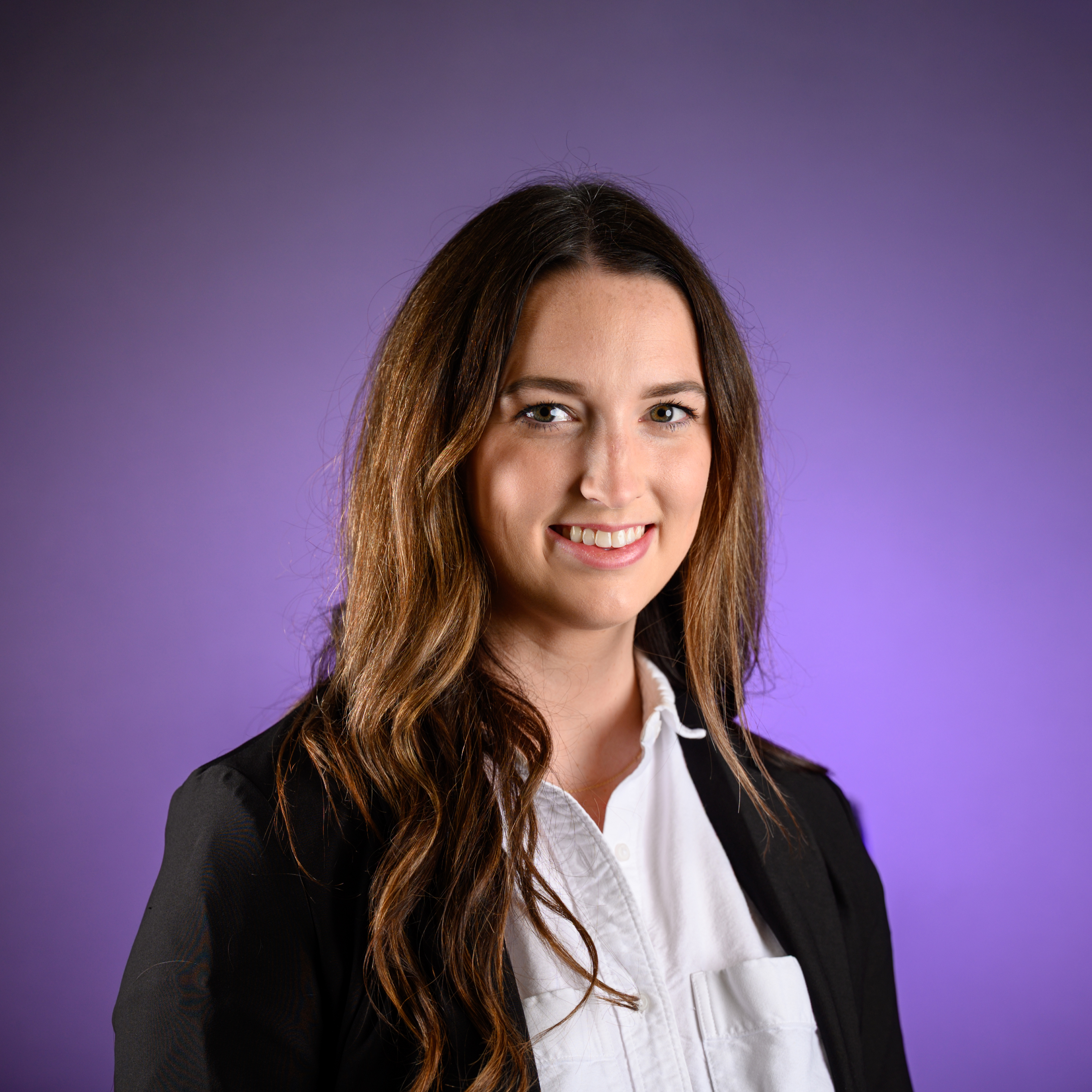Head-and-shoulders studio portrait of an adult with long wavy hair worn down, dressed in a black blazer over a white collared shirt, photographed against a smooth purple background.