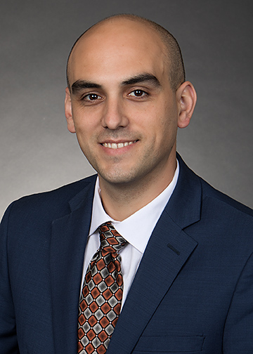 Professional headshot of a person wearing a blue suit jacket, white dress shirt, and a patterned orange-and-brown tie. The portrait is cropped from the chest up, with the subject facing slightly toward the camera against a dark gray studio background. The image is evenly lit and has a formal, business-style appearance suitable for a professional profile or staff directory.